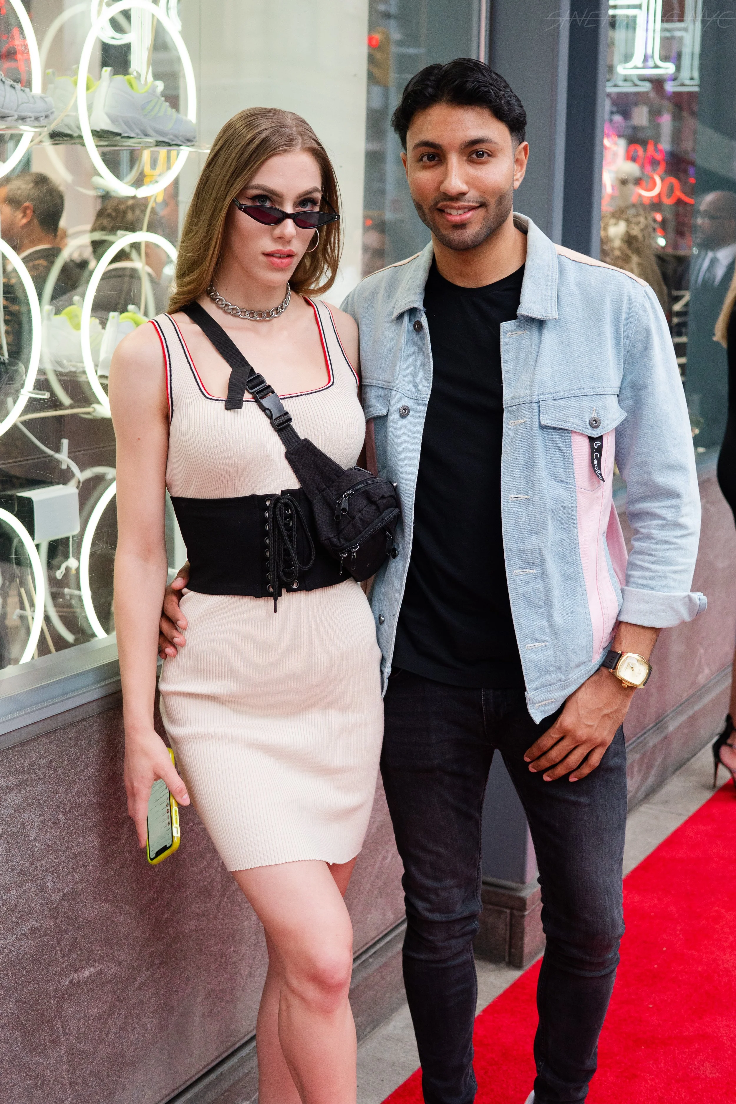 A young woman and man posing on a city sidewalk outside a storefront with shoes in the window. The woman wears a beige dress, sunglasses, a black waist bag, and holds a phone. The man wears a light denim jacket, black shirt, and a gold watch.