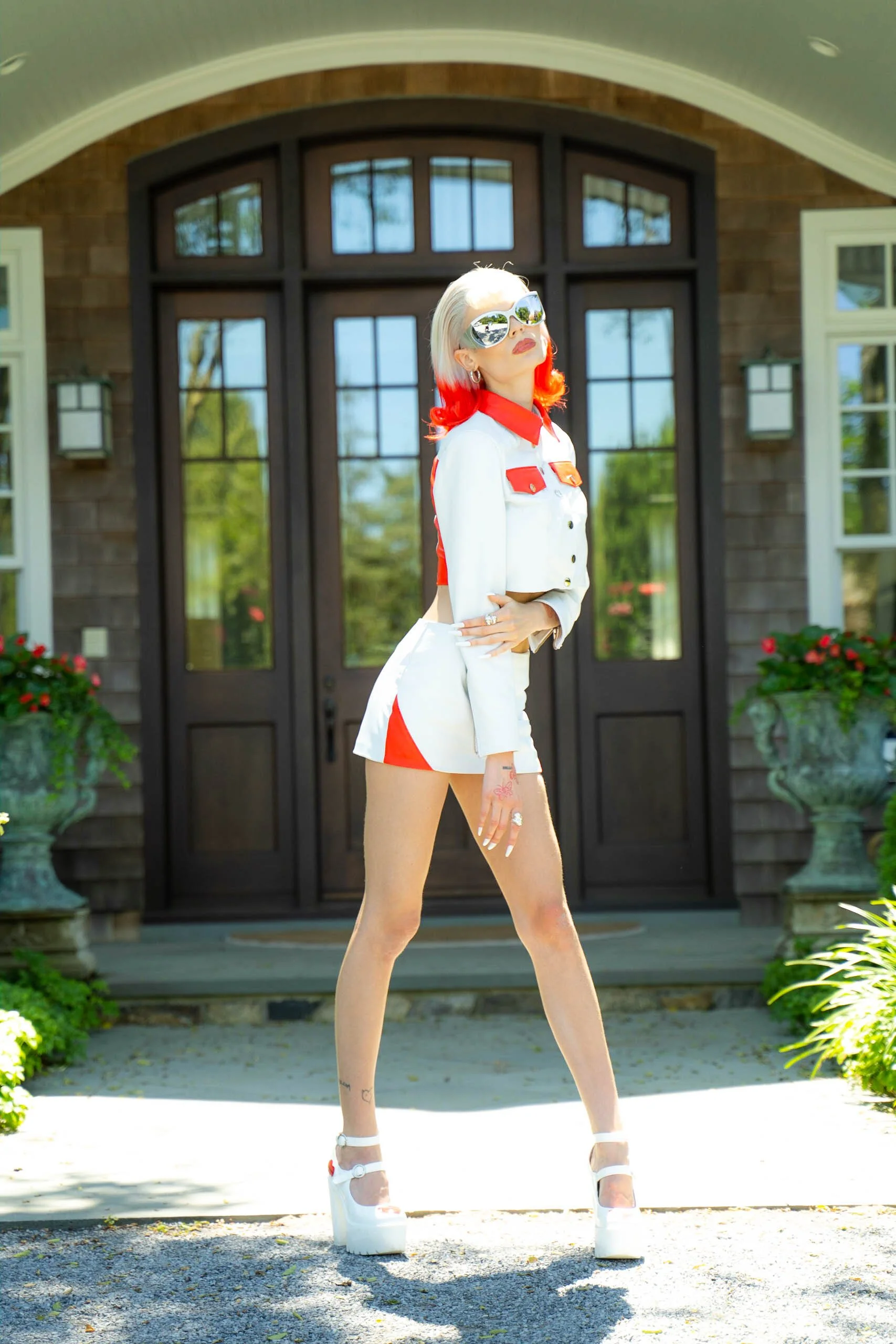 Woman with red and white hair wearing reflective sunglasses, a white and red jacket, matching white and red shorts, and white platform heels standing in front of a house with large glass door and potted plants.