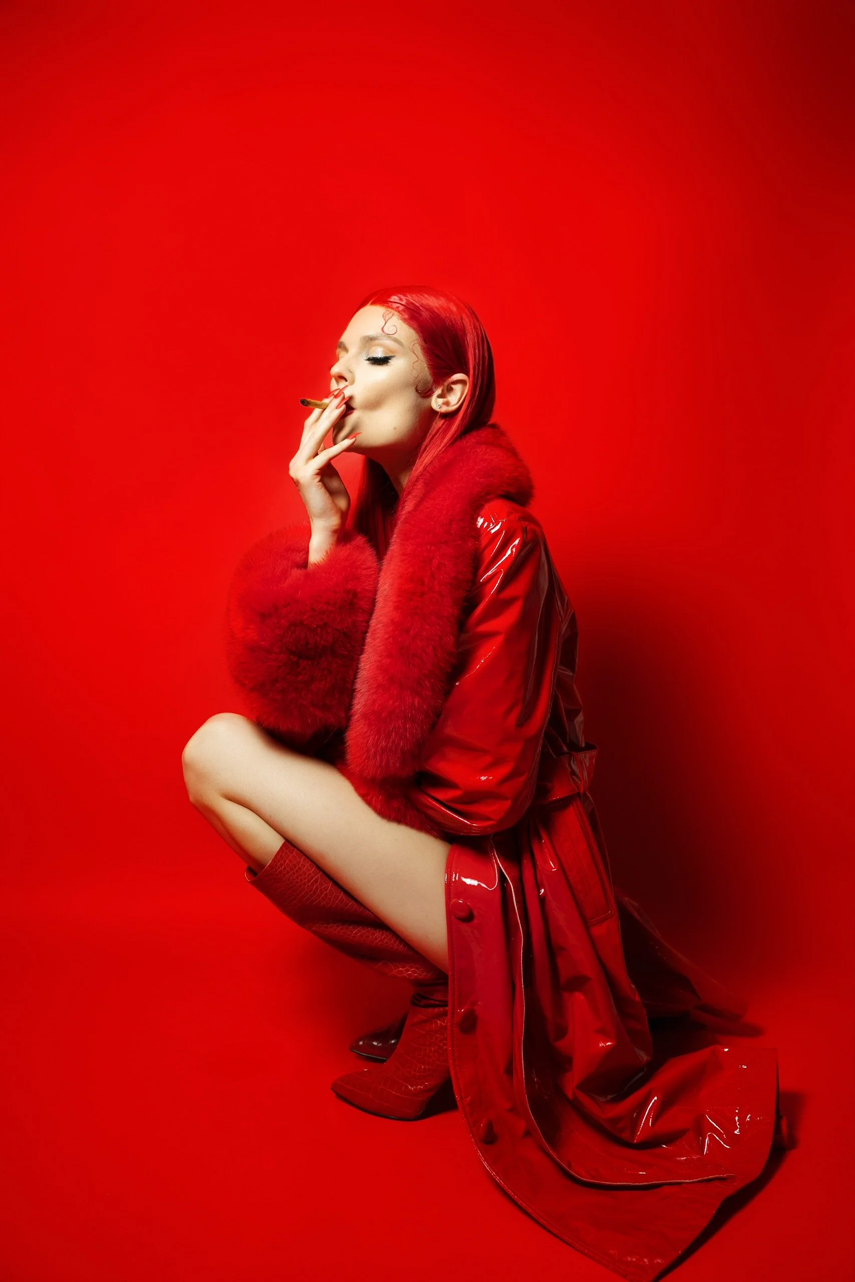 A woman with bright red hair, wearing a red fur jacket, red shiny coat, and red boots, posed against a red background, smoking a cigarette.