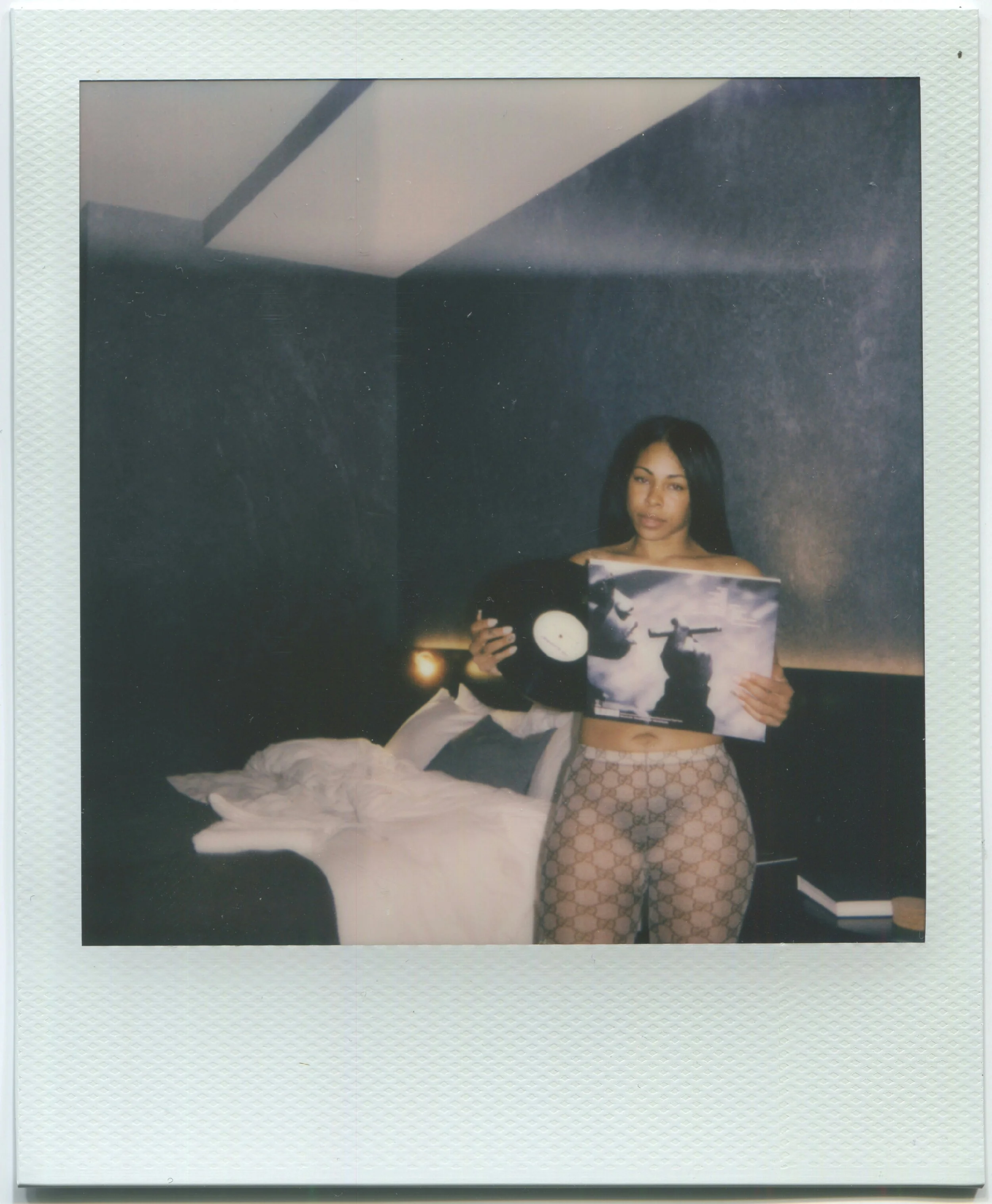 A woman with long black hair wearing Gucci patterned pants holding vinyl records and a vinyl cover in a dimly lit hotel room with a bed and dark textured wall in the background.