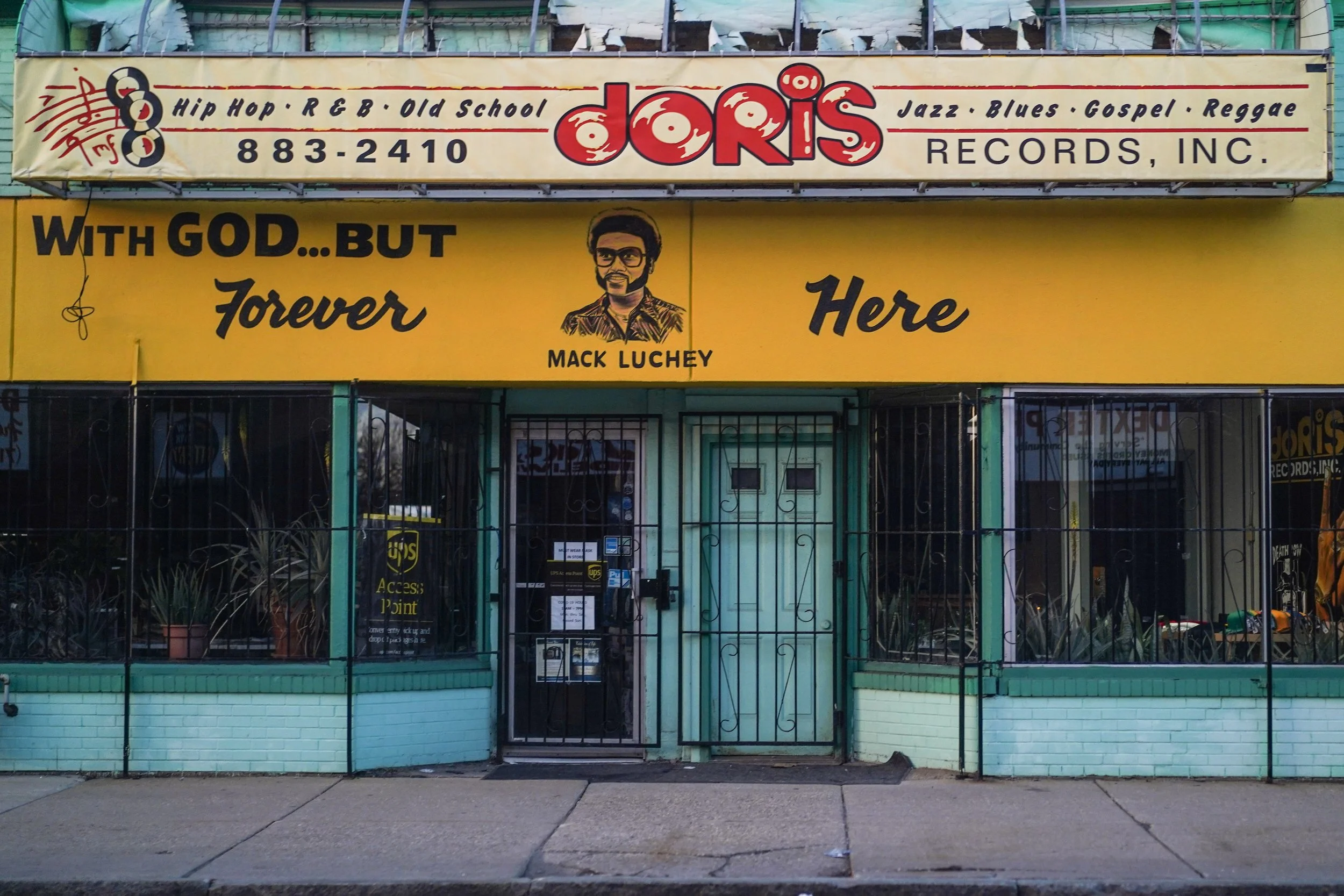  storefront with yellow and blue exterior, sign reading 'Doris Records, Inc.' with genres like hip hop and jazz, a portrait of Mack Luchey, and a quote 'With God... But Forever Here'.