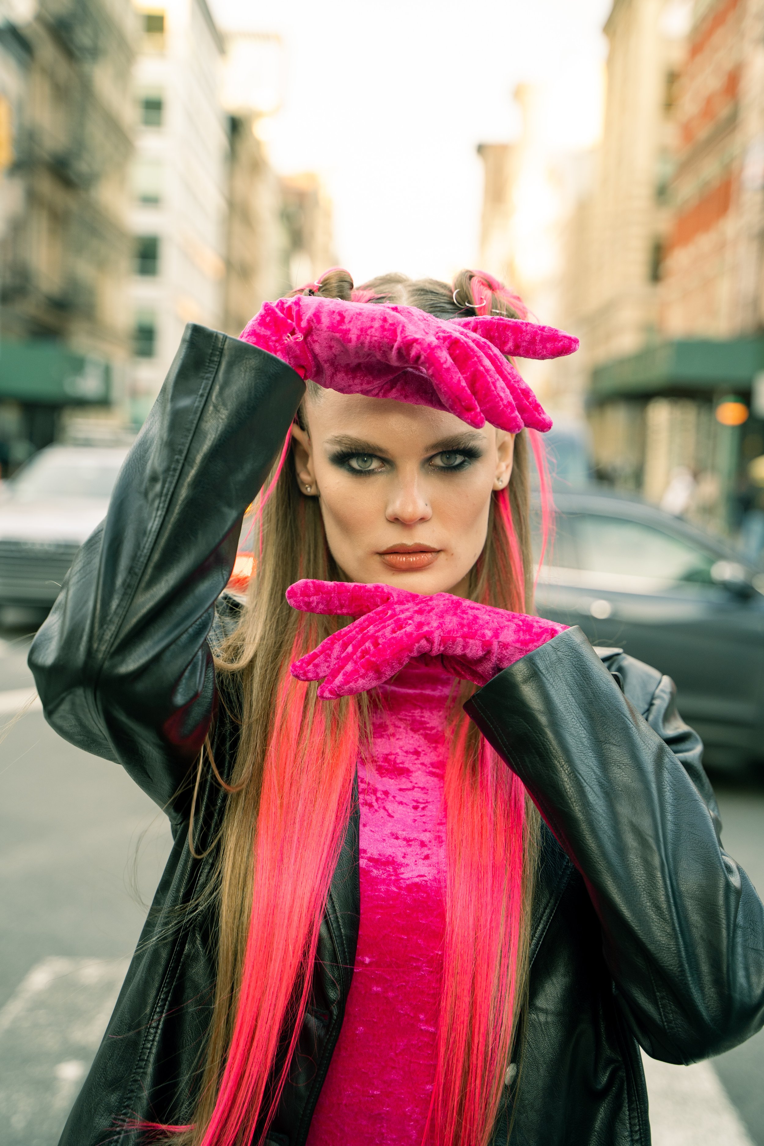 A woman with long pink hair and vibrant makeup poses in the street, wearing black leather jacket, pink velvet gloves, and a pink top, with city buildings in the background.