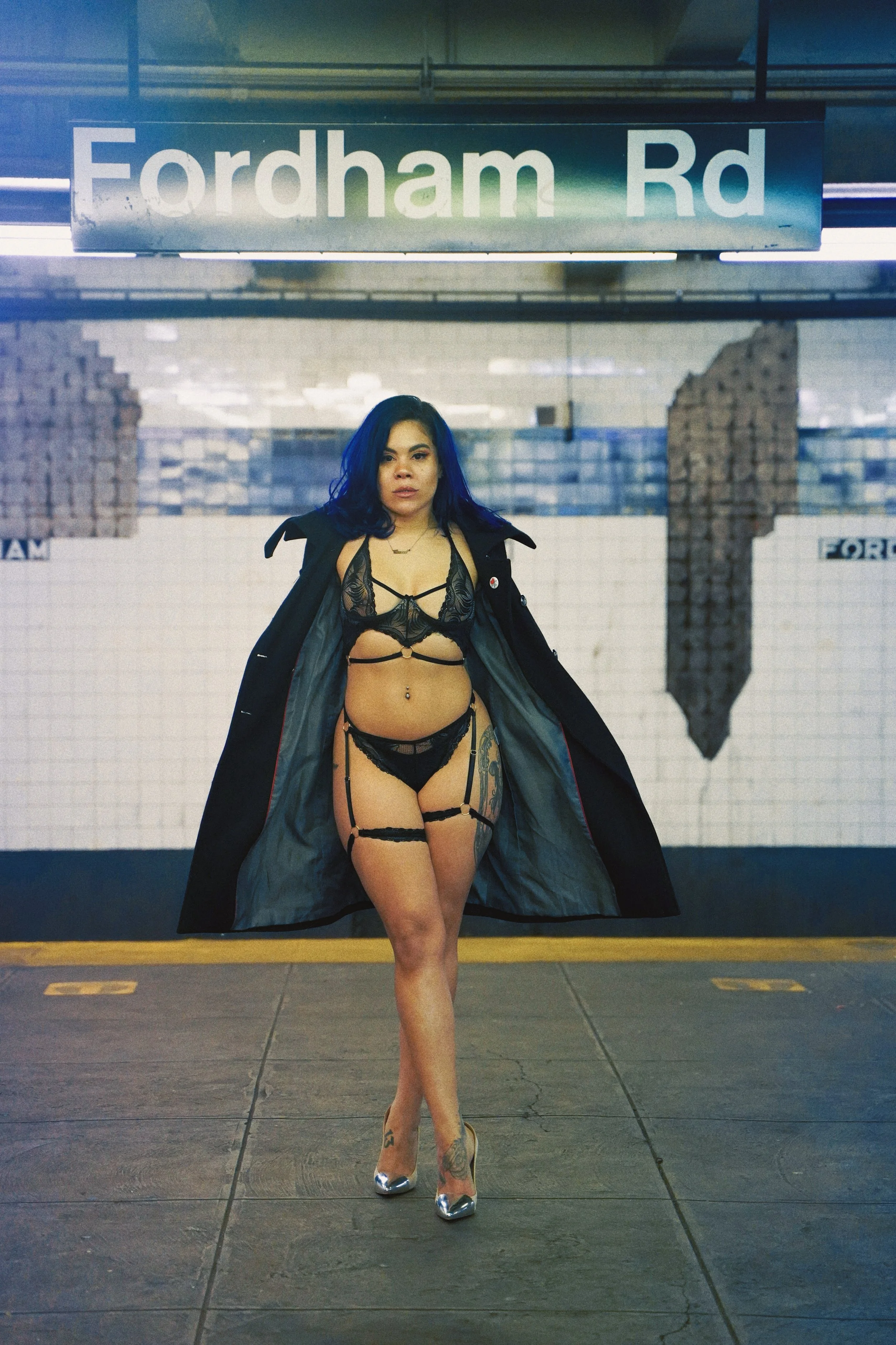Woman in black lingerie and stockings standing under Fordham Road sign in subway station.