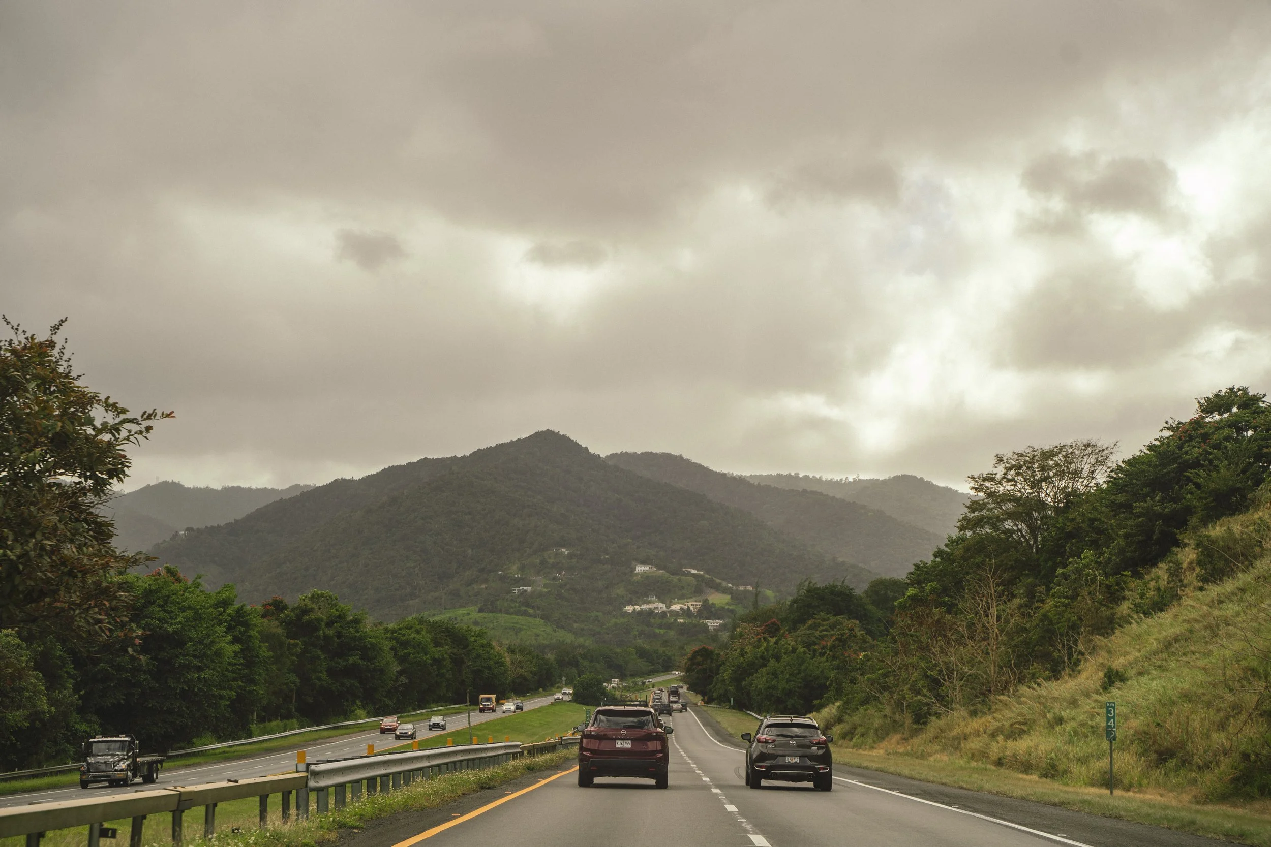 A highway with several cars driving in both directions, surrounded by green hills and mountains under a cloudy sky.