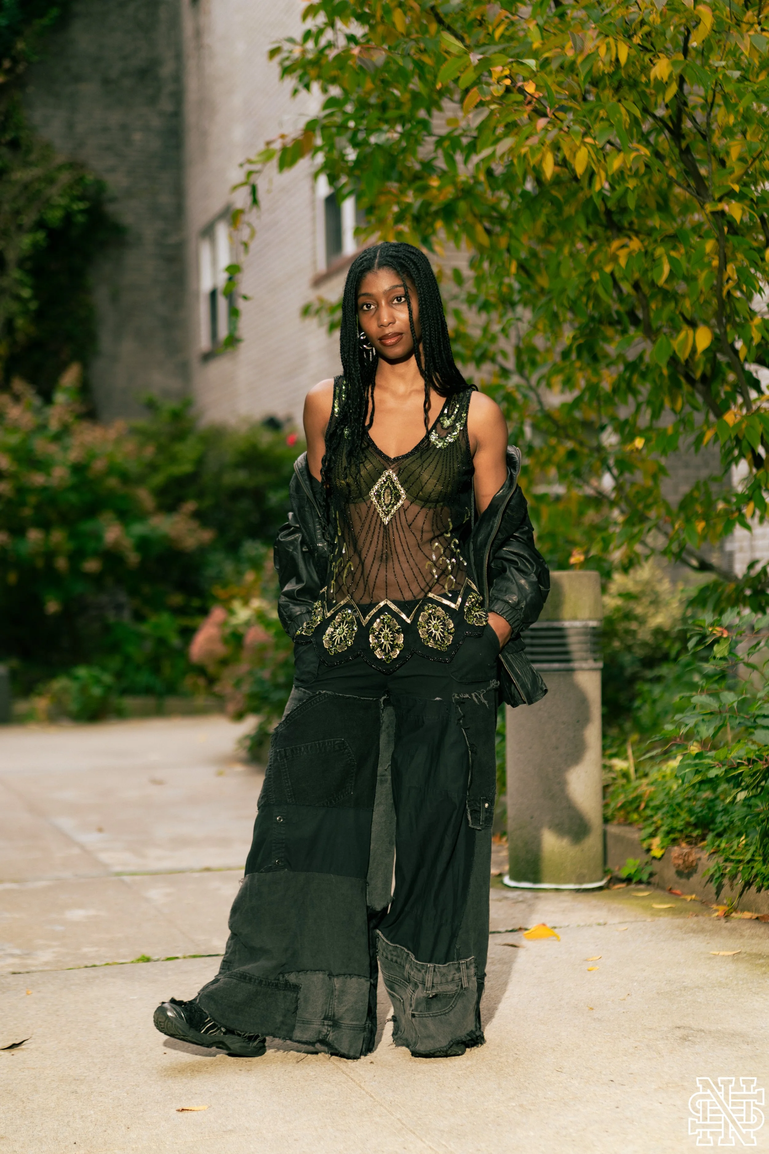 A woman with braided black hair, wearing a sheer black top with intricate gold embroidery, black wide-leg pants with multiple patches, and a black leather jacket draped over her shoulders, standing outdoors on a sidewalk next to a tree with green and