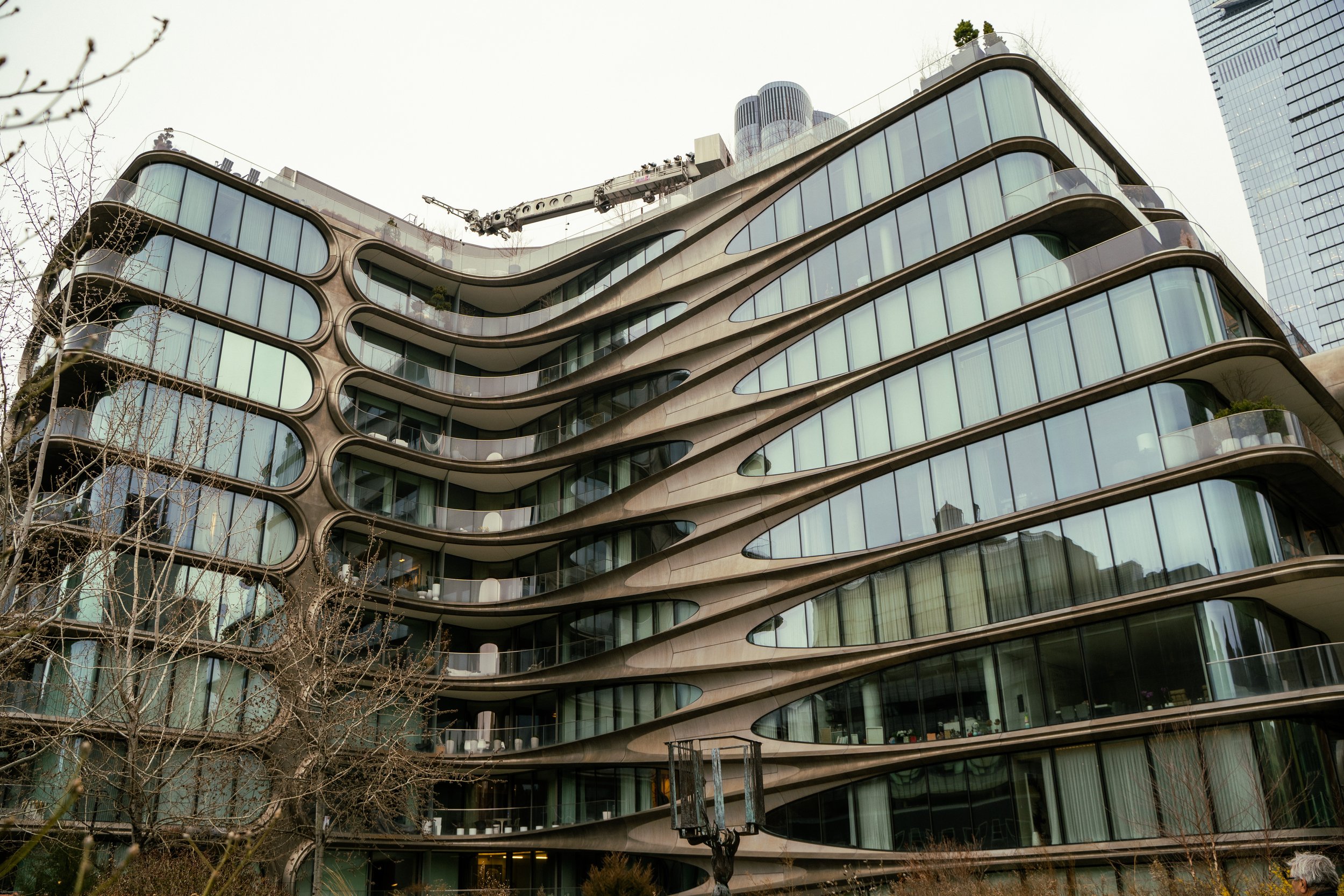 Modern multi-story building with curved glass balconies and large windows, set against a cloudy sky, with leafless trees in the foreground.