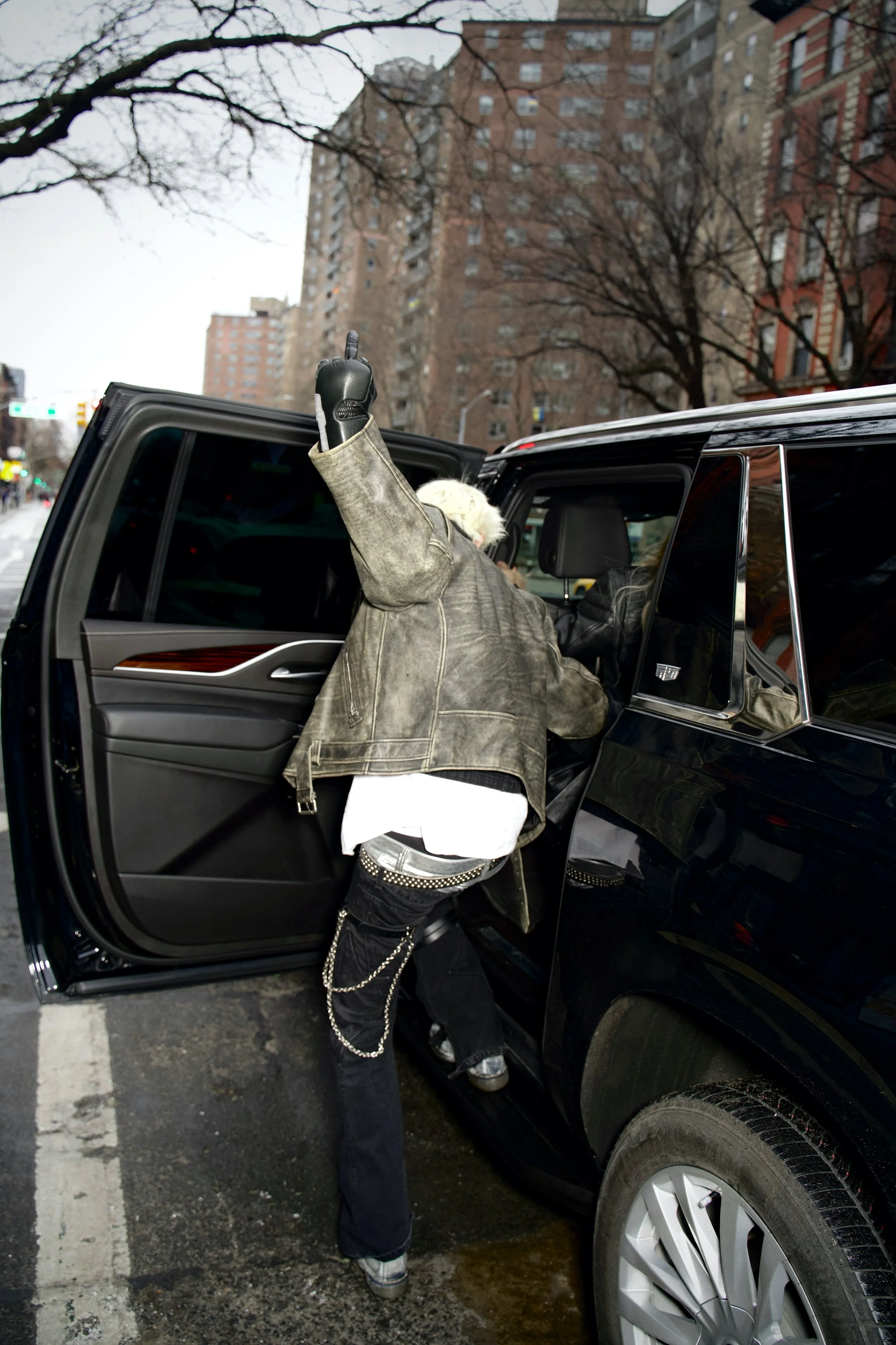 Person getting into a black SUV on a city street, with tall buildings and leafless trees in the background.