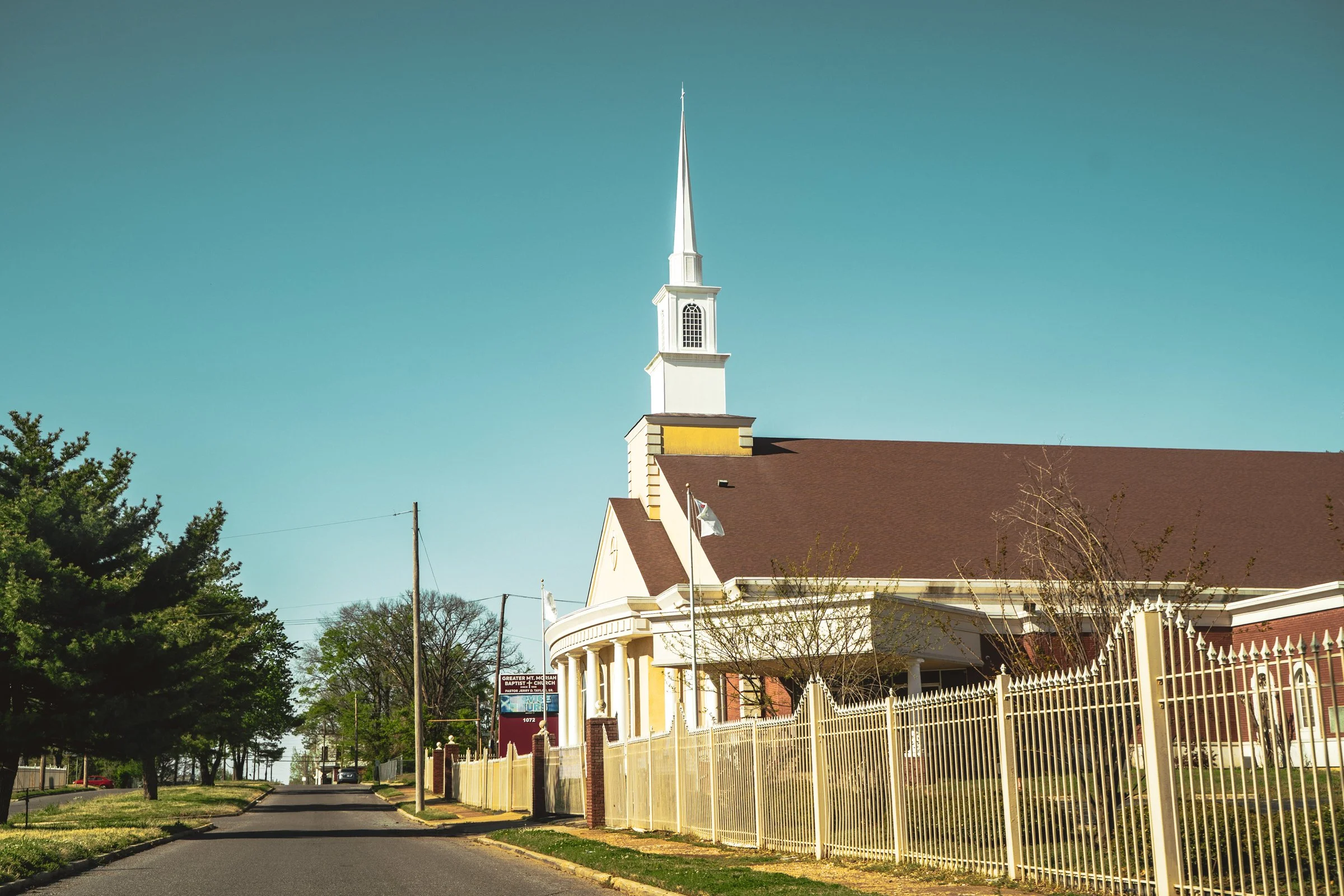 A white church with a tall steeple and a brown roof, surrounded by a white fence, located along a suburban street with trees and utility poles.