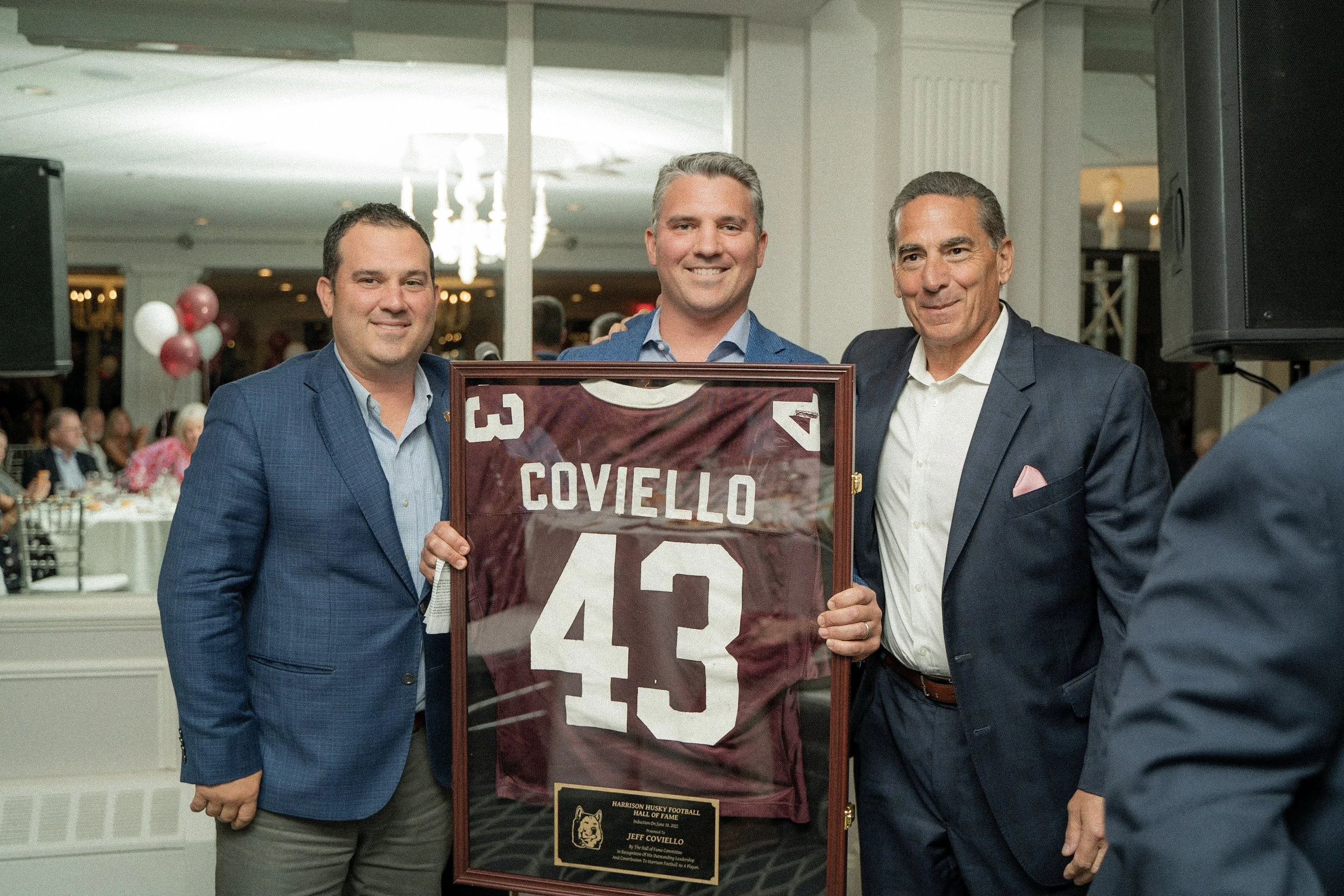 Three men in suits are standing inside a banquet hall, holding a framed football jersey with the name 'Coviello' and the number 43. They are smiling, and there are other guests seated at tables in the background.