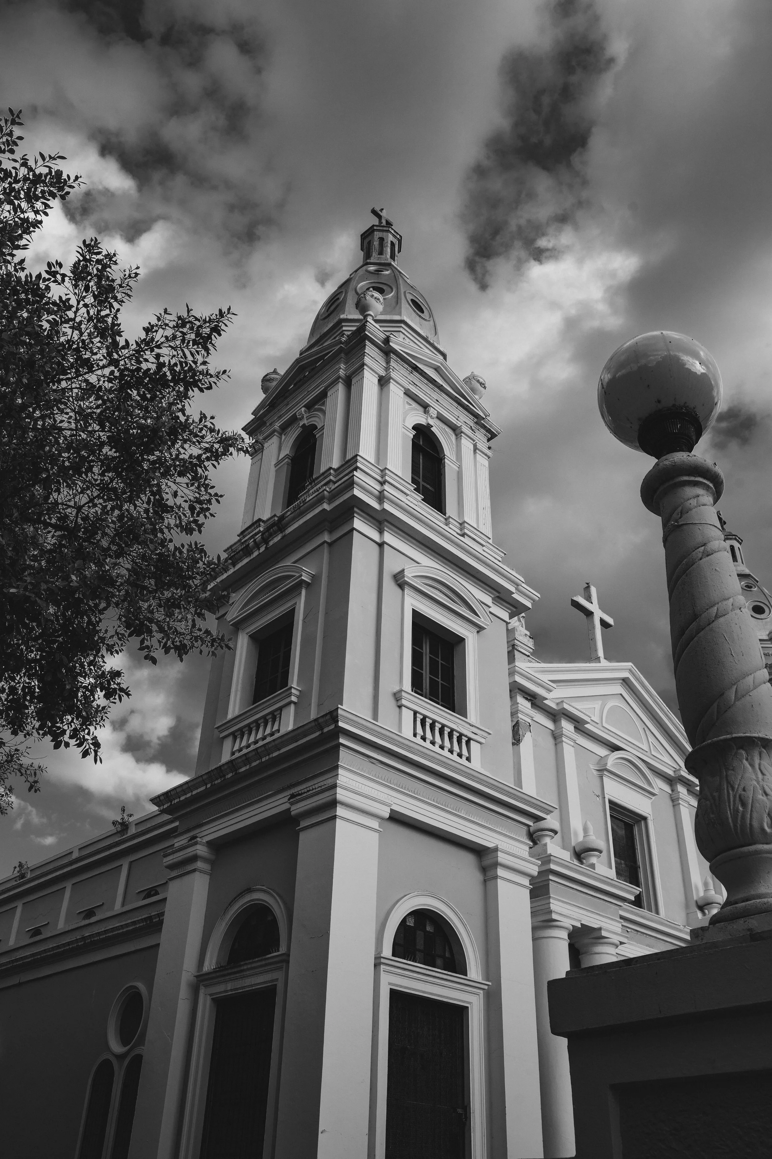 A black and white photo of a church with a tall steeple topped with a cross, decorative architectural details, arched windows, and a cloudy sky in the background.