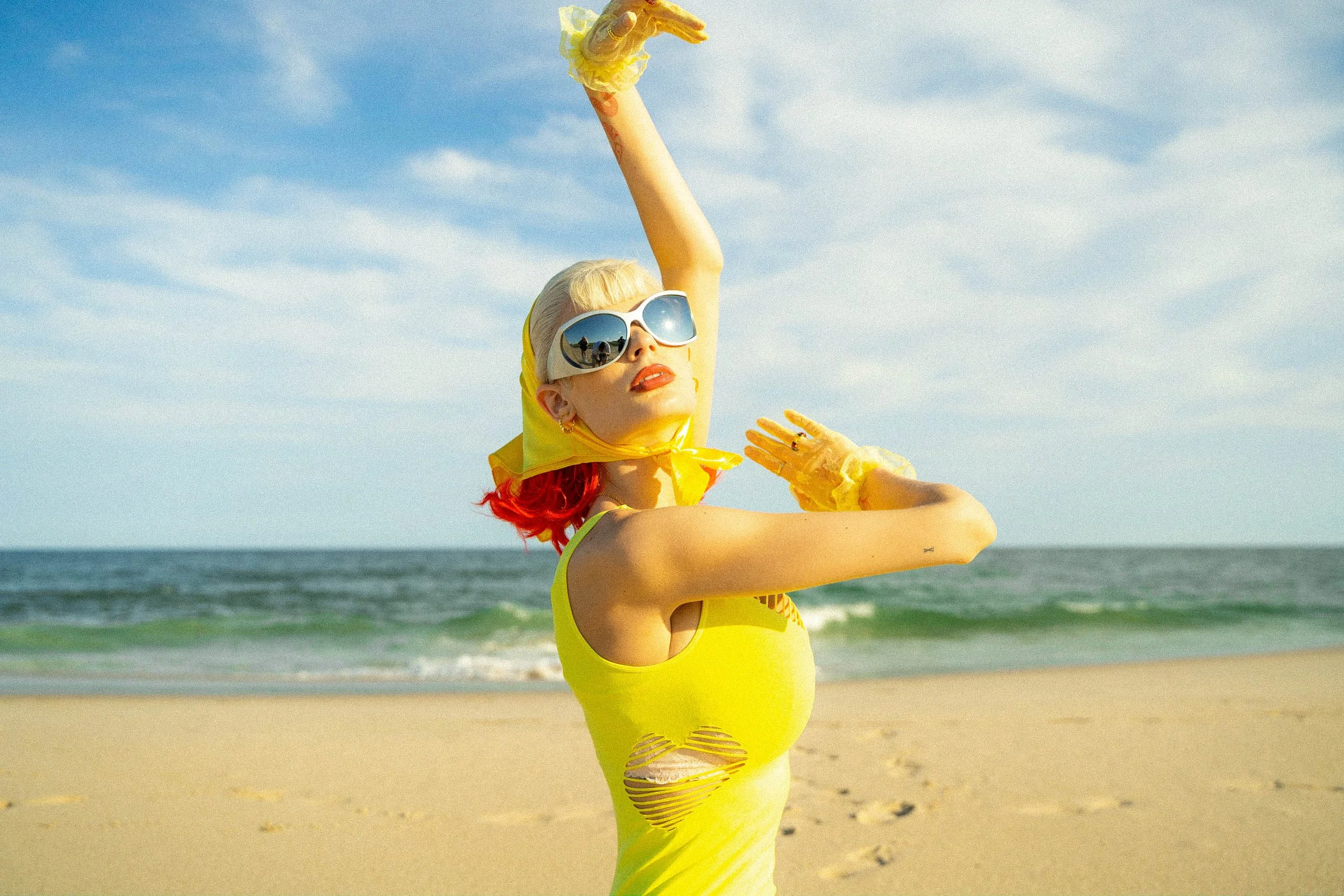 A woman with blonde hair and red tips, wearing large sunglasses, a yellow scarf, and a yellow dress with ripped details, standing on a beach. She is wearing yellow gloves and striking a pose with one arm raised and the other bent. The background show