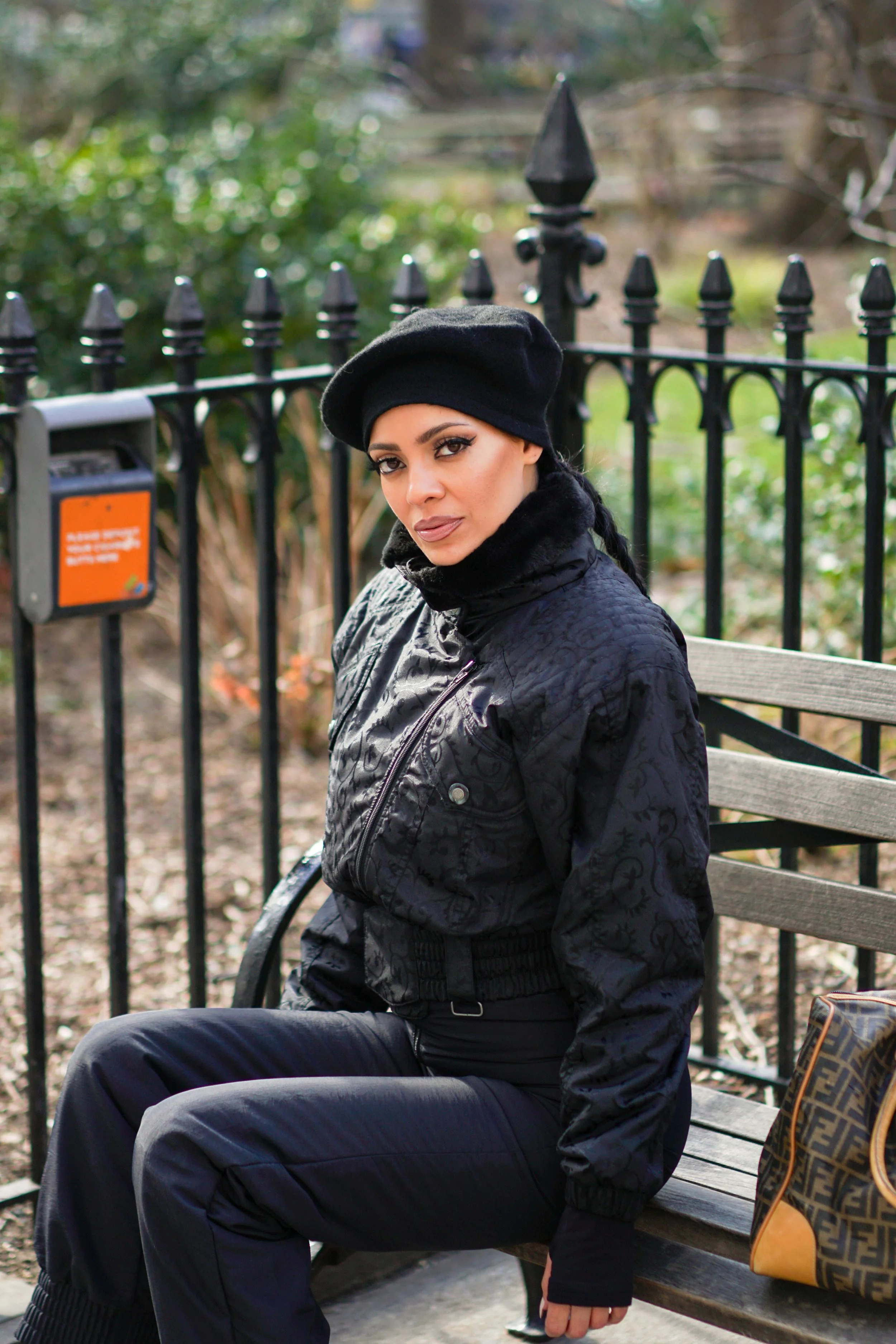 A woman sitting on a park bench wearing a black beret, black jacket, and black pants. She has dark hair and makeup, with a brown patterned handbag next to her. The background features a black wrought iron fence and greenery.