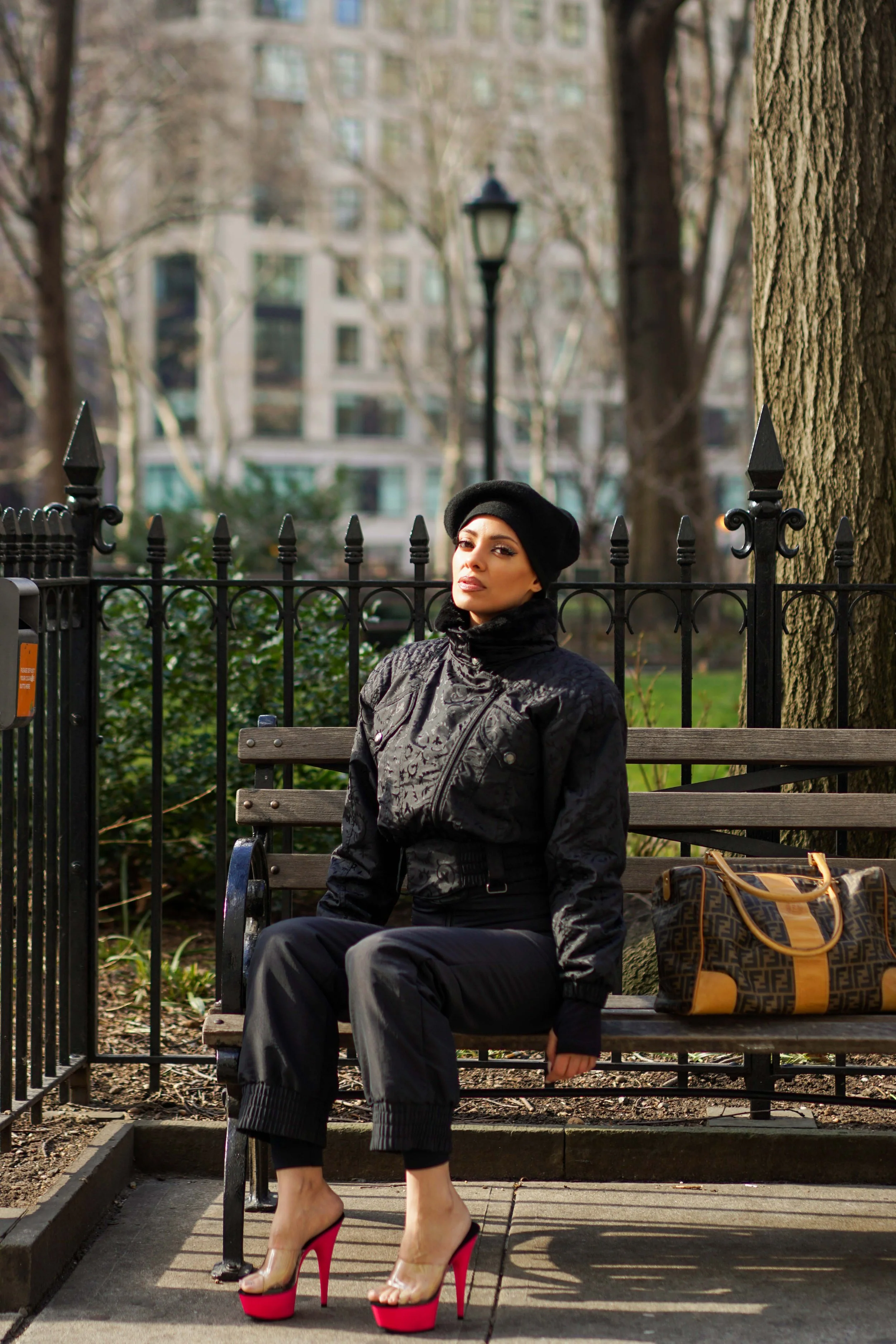 A woman sitting on a park bench with a handbag beside her, wearing a black beret, black jacket, black pants, and high-heeled shoes, in an urban park setting with trees and buildings in the background.