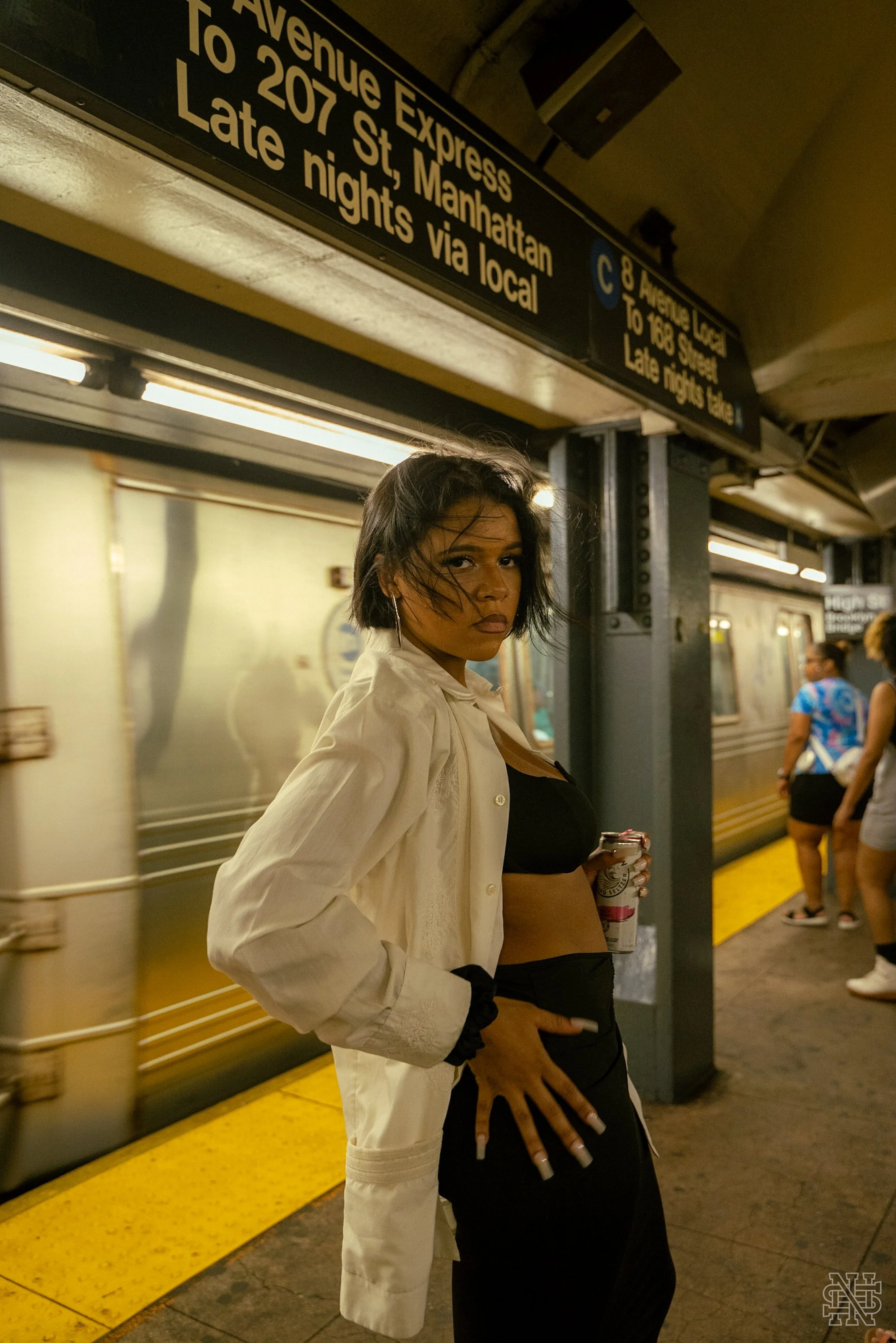 A woman with short black hair, wearing a white shirt over a black outfit, standing on a subway platform holding a drink, with groups of people in the background and a train arriving or departing.