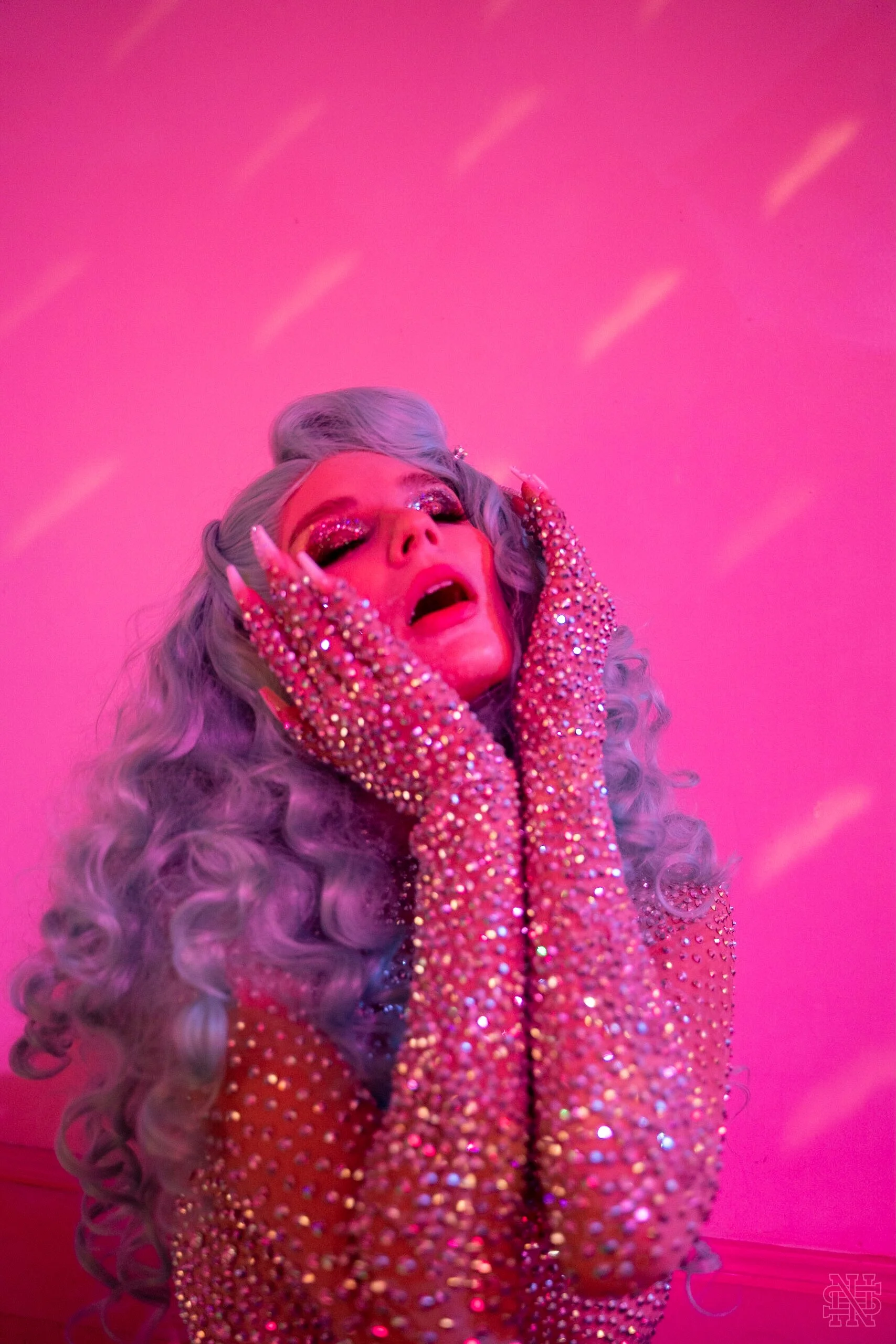 A person with long, wavy, grey hair and glittery eye makeup posing with hands on their face, wearing a rose-colored, sparkly outfit, against a pink background.