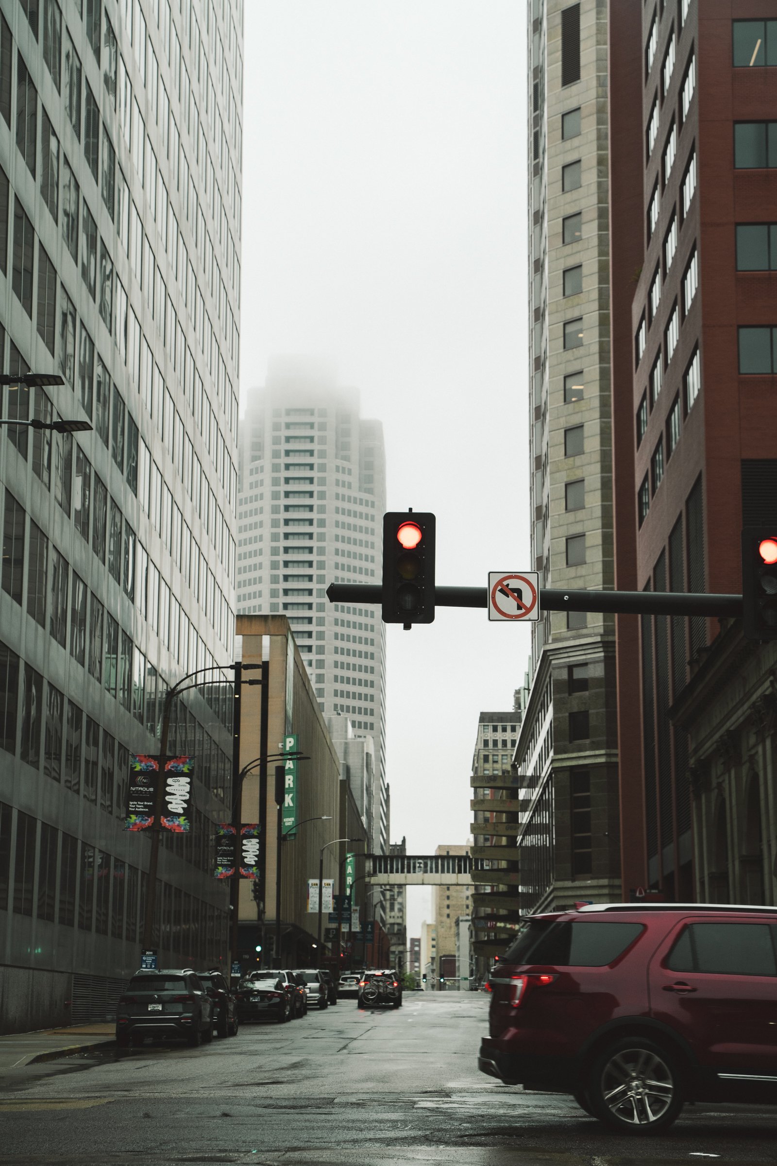 A city street on a rainy day with tall buildings, parked cars, and a traffic light showing red.
