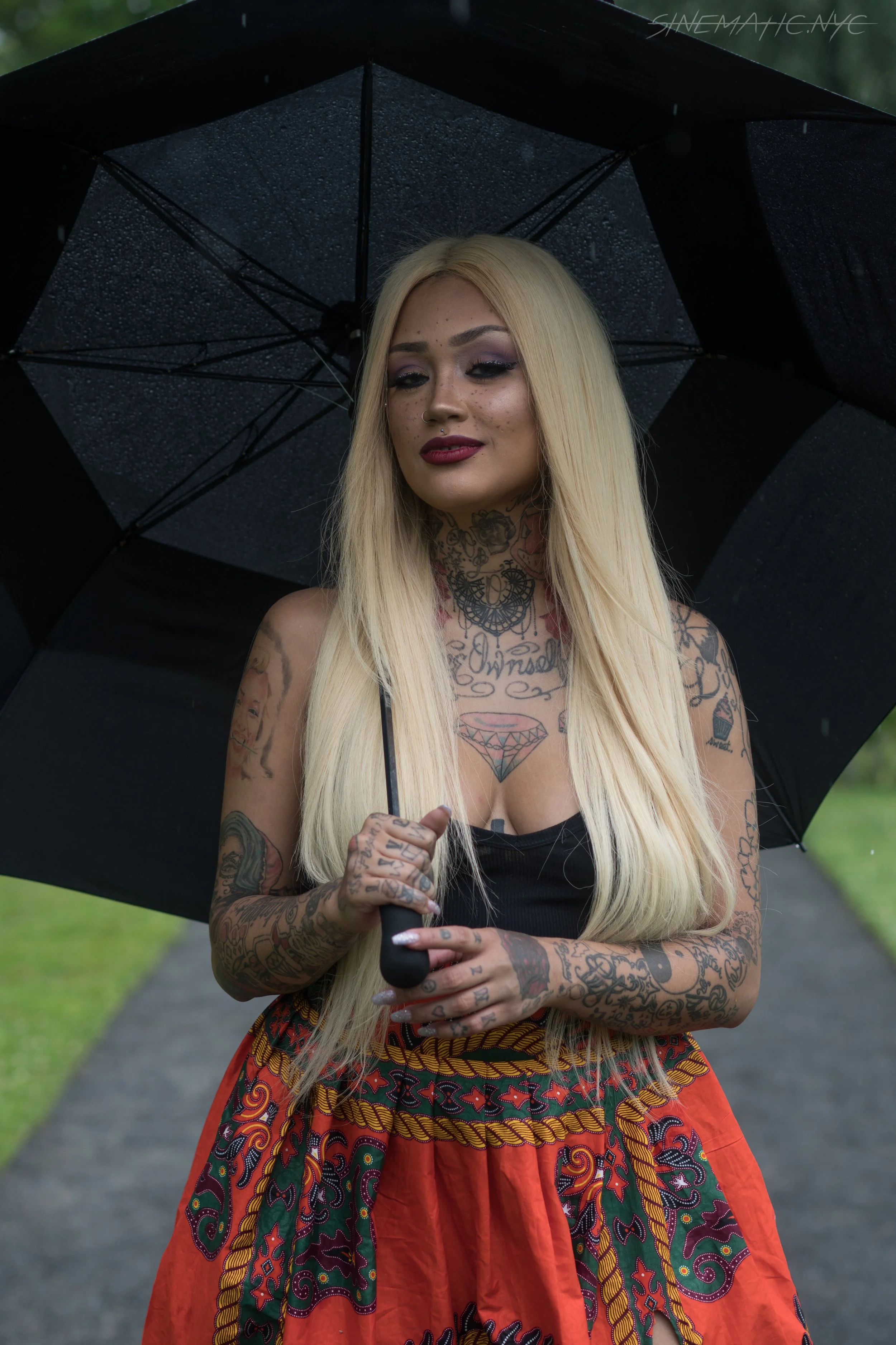 A woman with long blonde hair, tattoos on her arms and chest, and dark makeup holds a black umbrella outdoors in the rain.