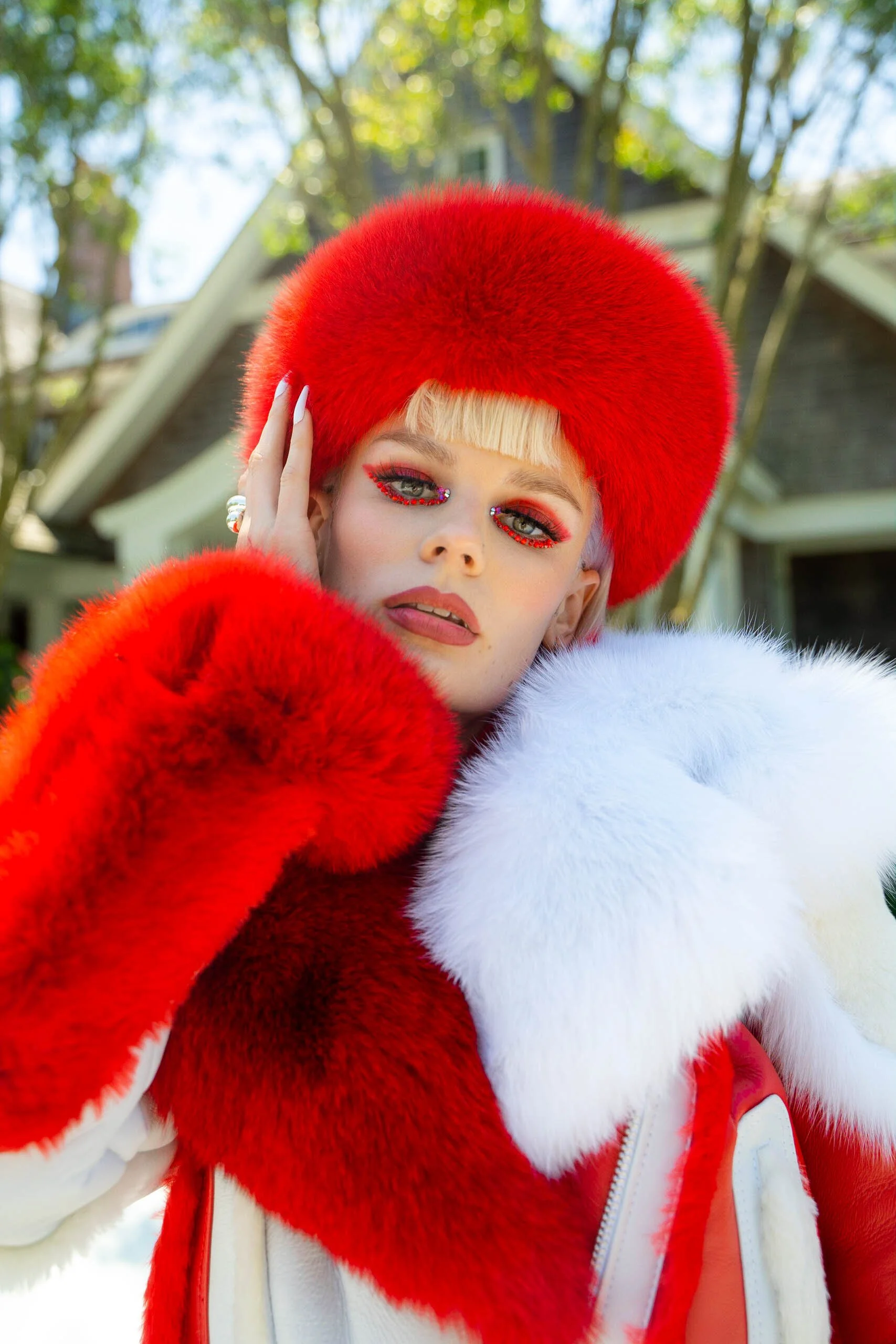 A person with platinum blonde hair wearing a large red fur hat, red fur gloves, and a white and red fur coat posing outdoors with trees and a house in the background.