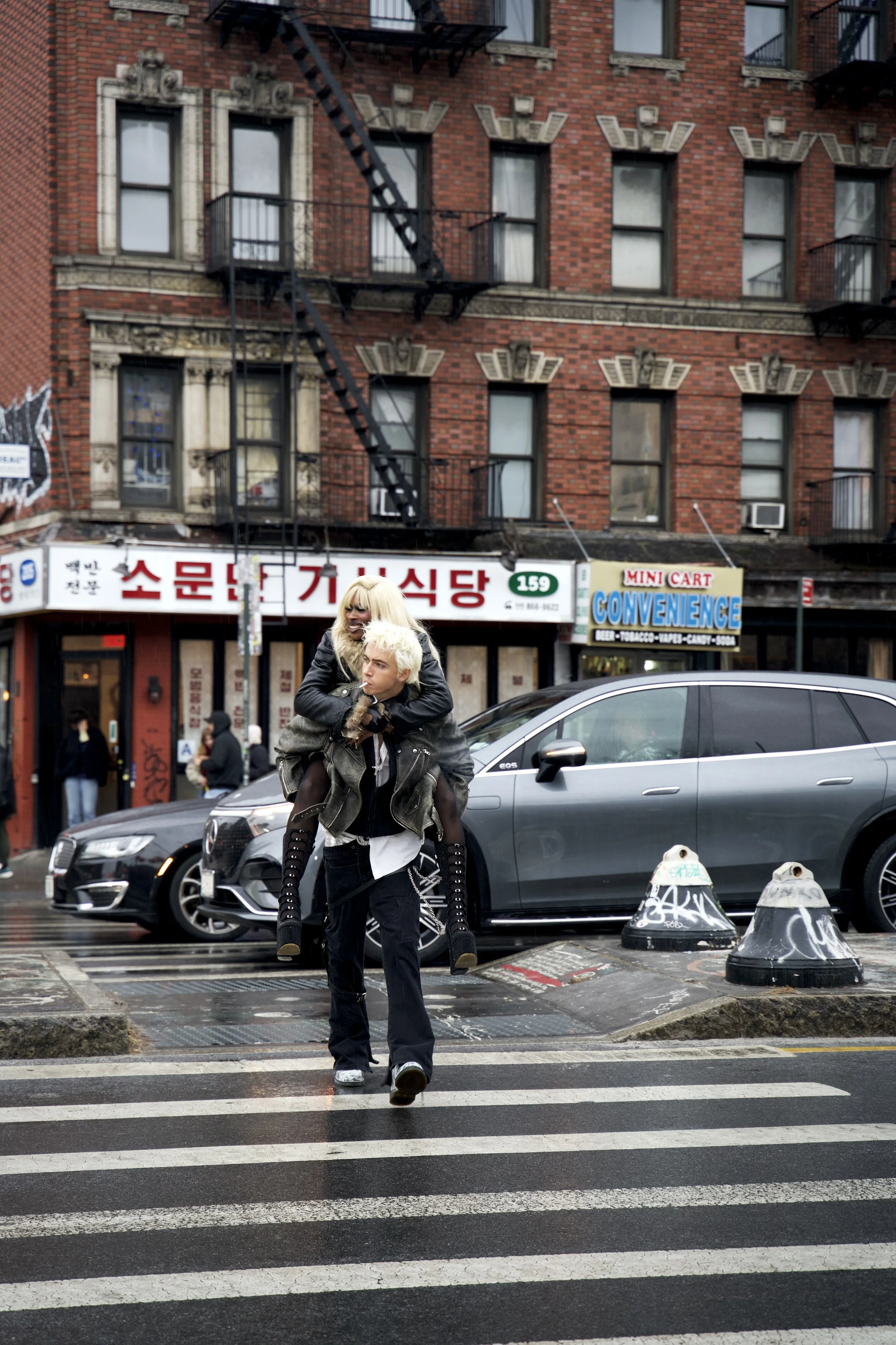 Two young women with platinum blonde hair, wearing dark clothing, are crossing a city street with cars parked along the curb and brick buildings in the background.