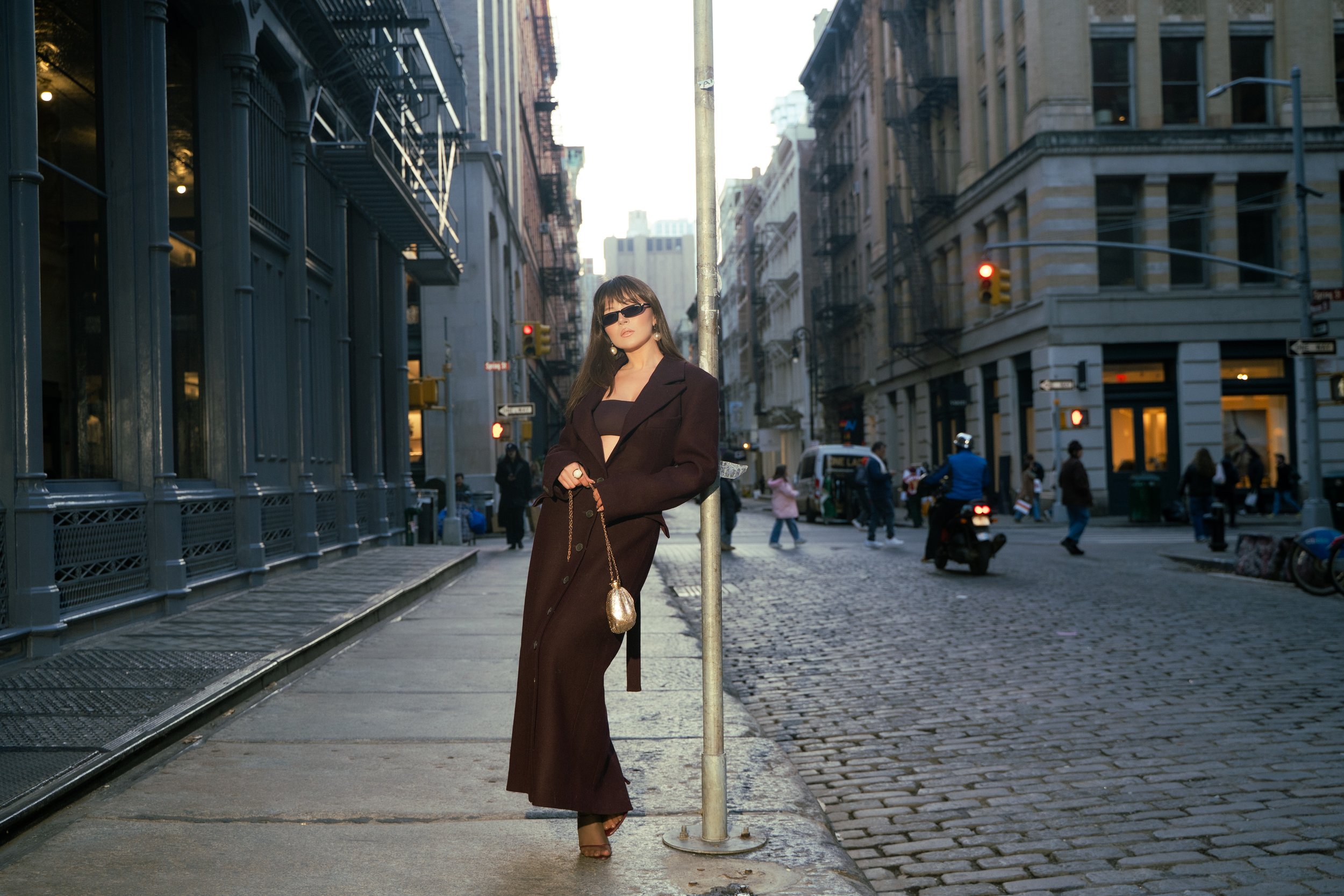 A woman in a dark brown coat, black top, and sunglasses standing on a city sidewalk leaning against a street pole, with buildings and pedestrians in the background.