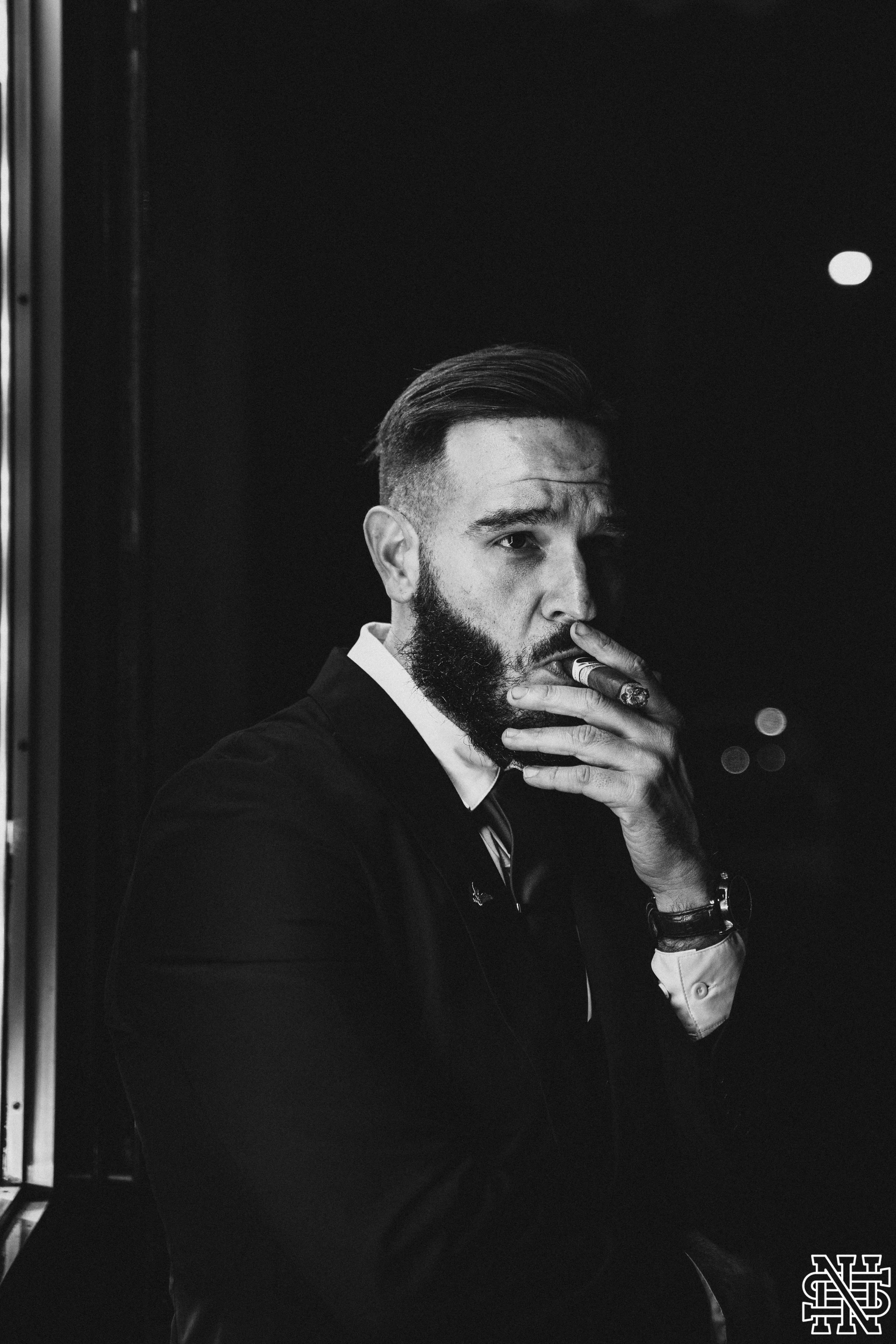 Black and white photo of a man with a beard smoking a cigar, wearing a suit, posed thoughtfully with one hand near his face, sitting near a window at night.