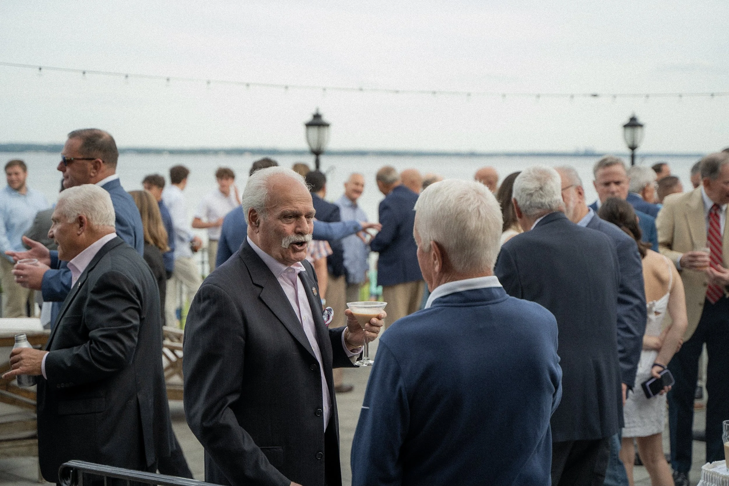 A group of people at an outdoor social event by the water, dressed in formal and semi-formal attire, with some holding drinks, and a view of the water and sky in the background.