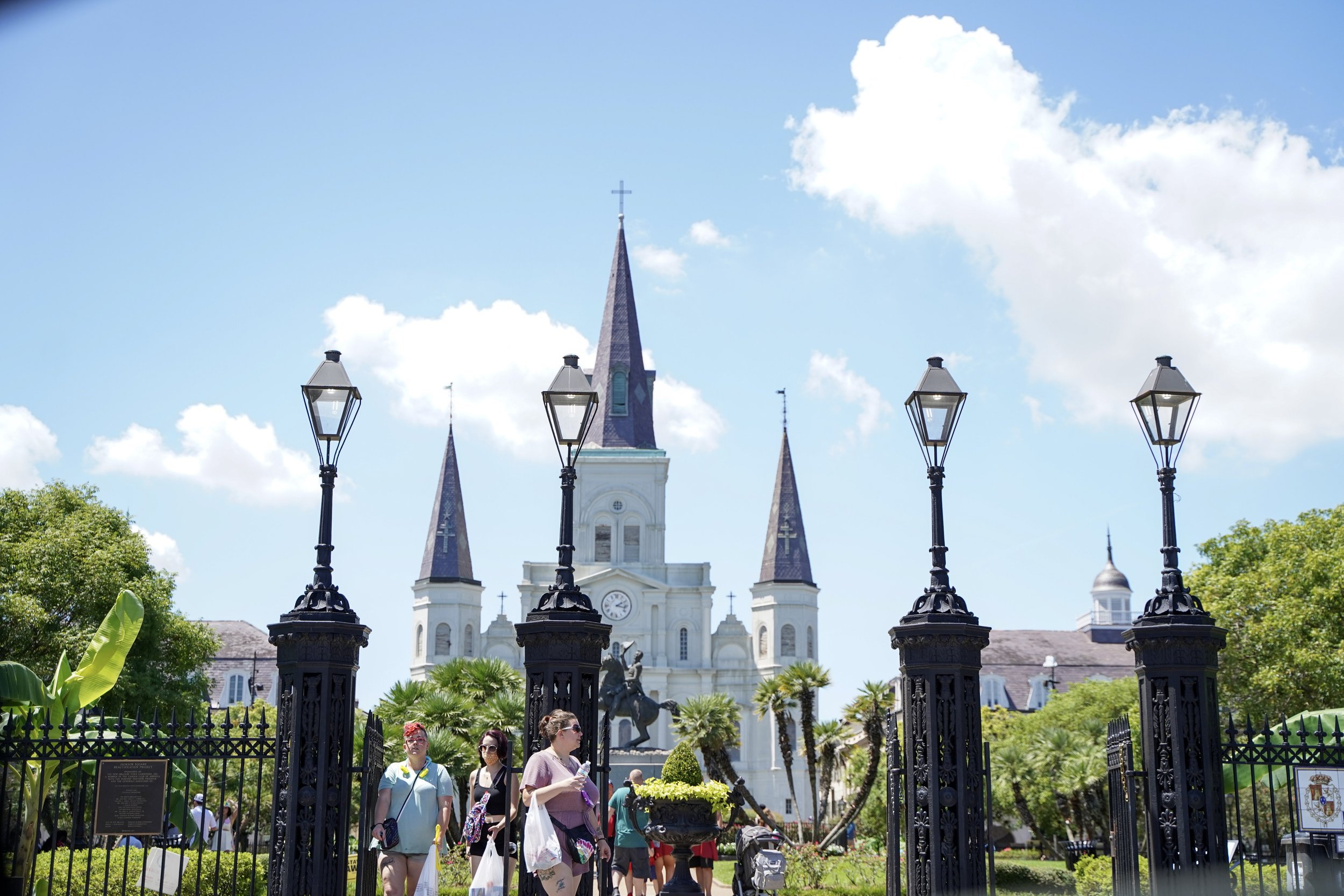 The image shows a white church with three steep purple-spire towers, set behind a black wrought-iron fence and with four street lamps at the entrance. People are walking in the garden area in front of the church, surrounded by lush green trees and pl