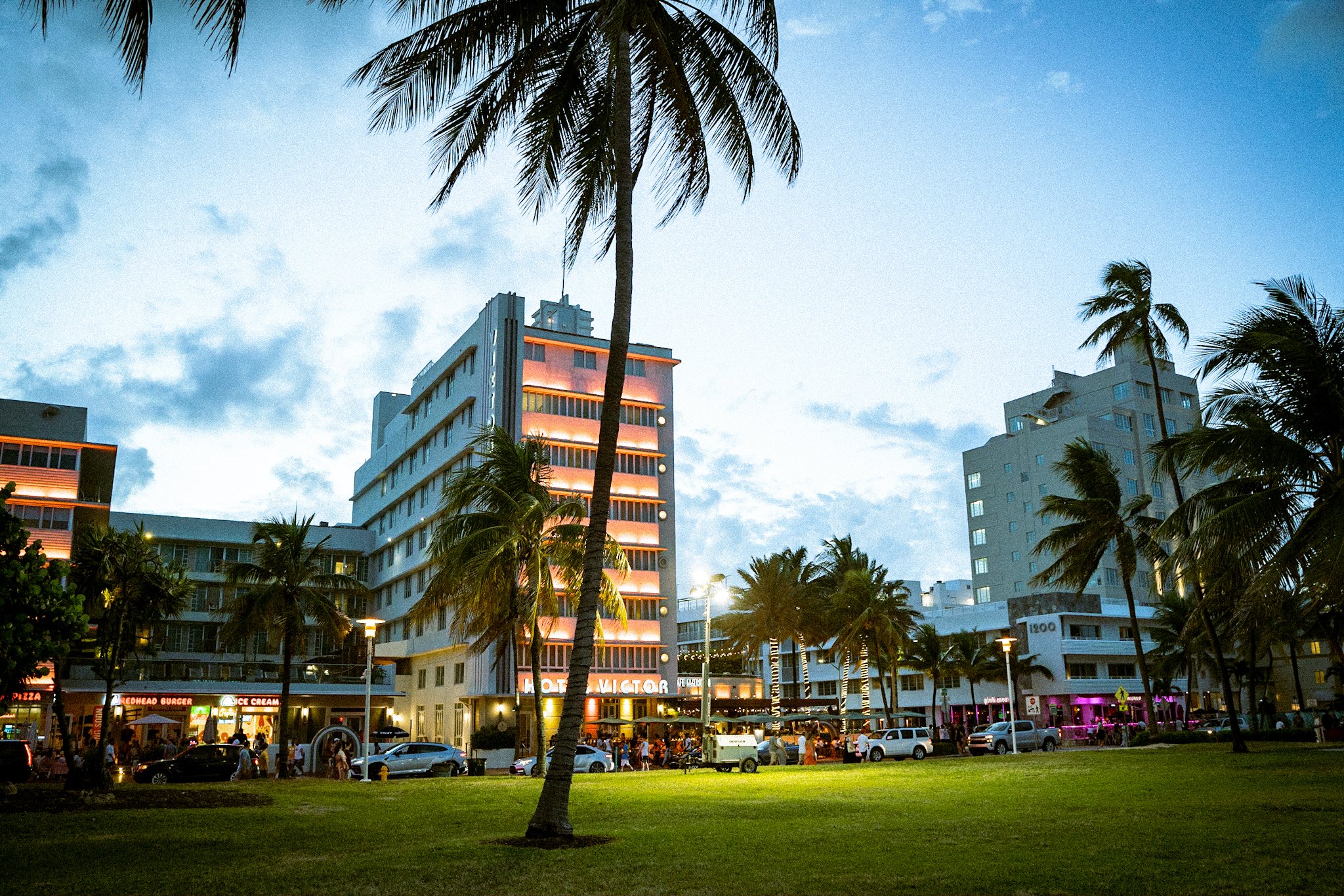 A Miami city street scene at dusk with tall buildings, palm trees, and people gathered outside of shops and restaurants.