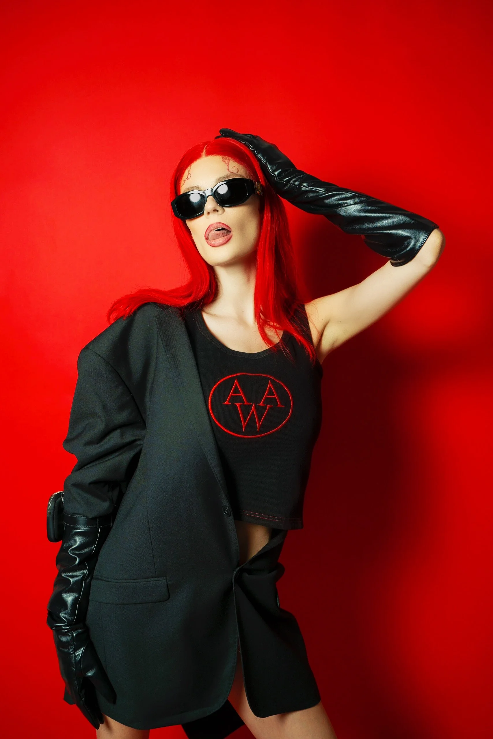 A woman with bright red hair wearing sunglasses, black gloves, a black crop top with red logo, and a black blazer standing against a red background.
