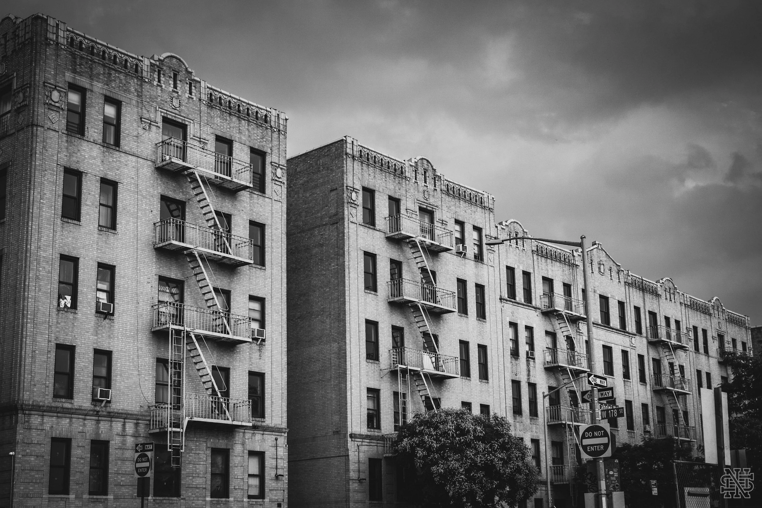 Black and white photo of multi-story brick apartment building with fire escapes on the front and street signs at the corner.