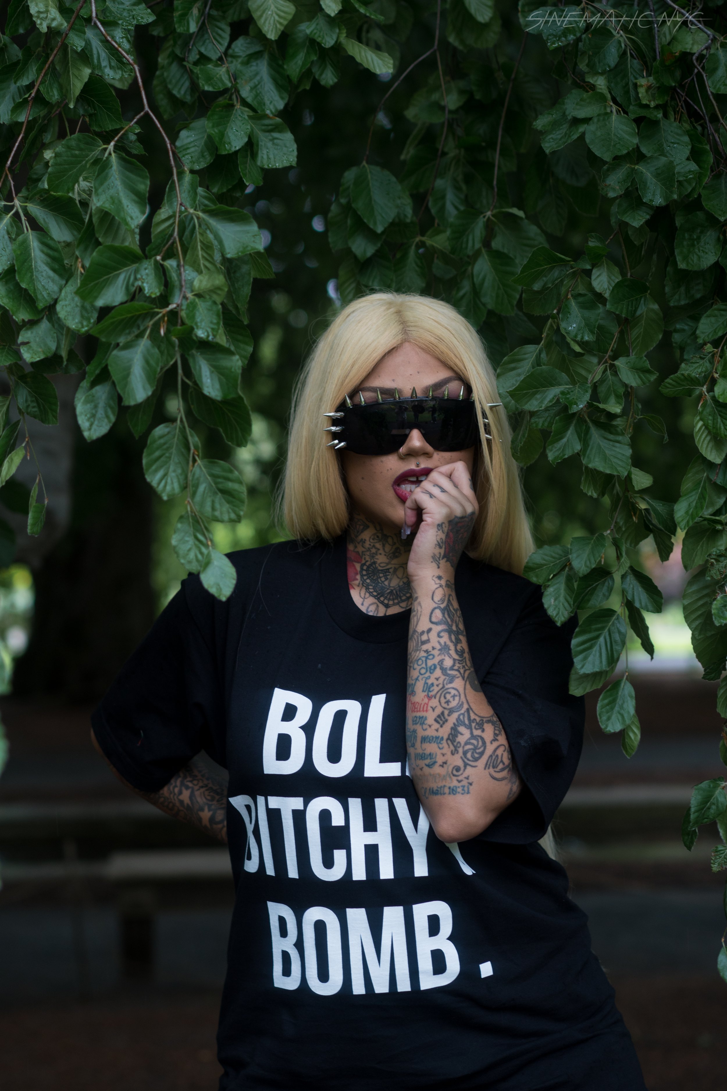 A woman with blonde hair wearing black sunglasses with metal spikes, a nose ring, and tattoos on her arm and neck, standing outdoors among green leaves. She is wearing a black T-shirt with white lettering and has her hand near her mouth.