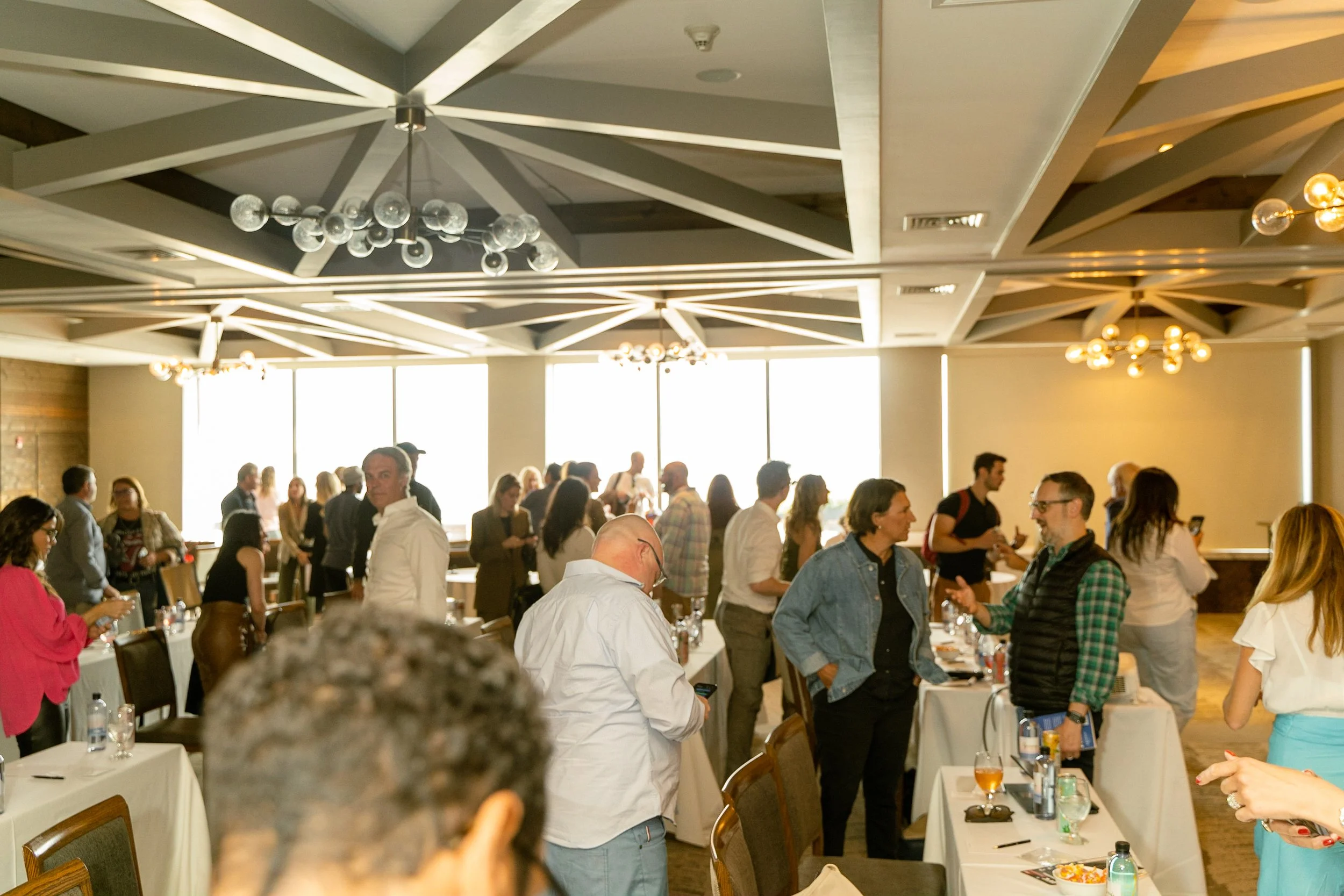 People gathered in a bright, modern conference room with tables, engaged in conversation, with large windows letting in sunlight, and decorative ceiling lights.
