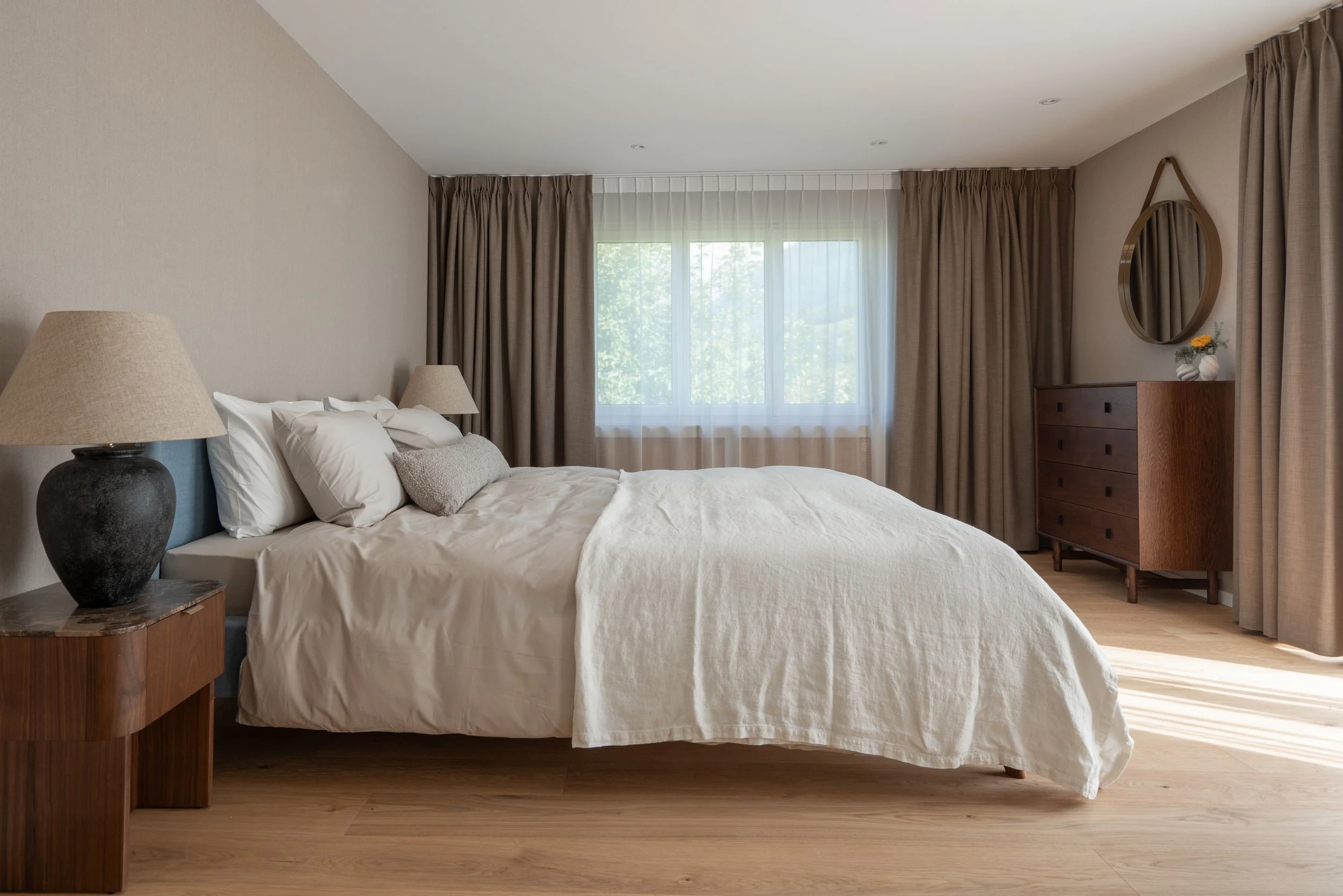 Master bedroom with linen-textured wallpaper, a soft blue upholstered headboard, neutral bedding, warm taupe curtains and a wooden chest of drawers, bringing together a warm and tactile palette of materials.