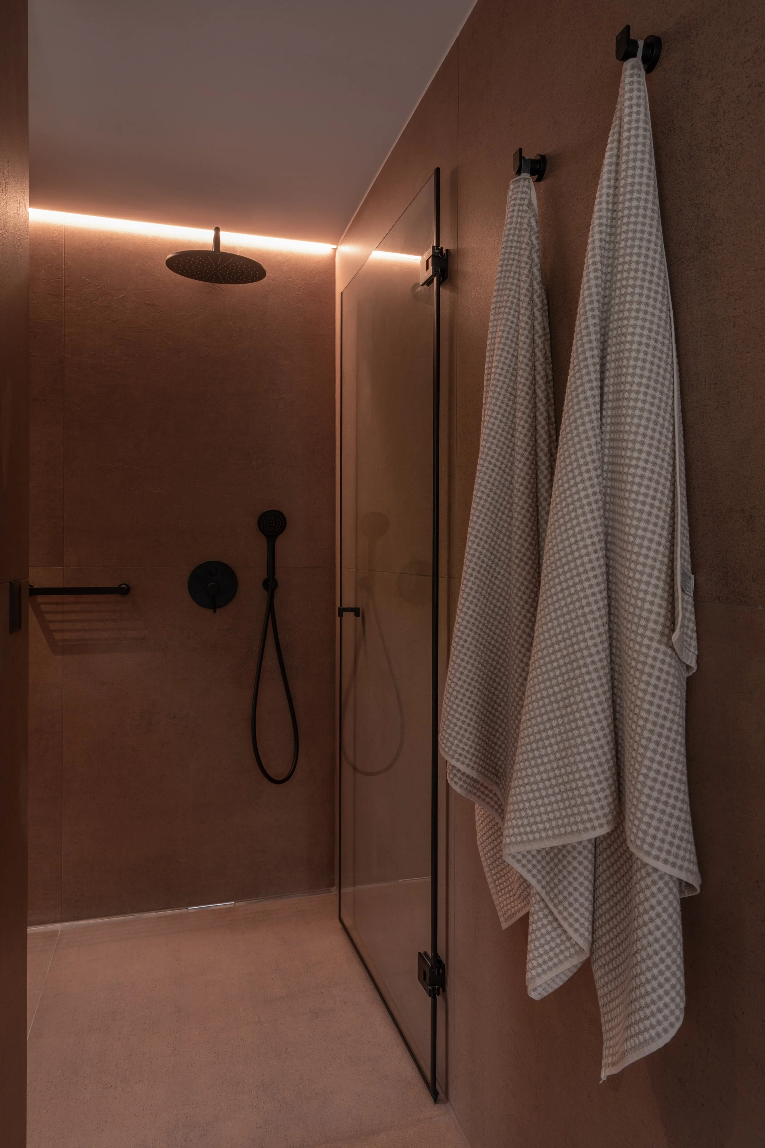 Walk-in shower finished in warm earthy-toned surfaces with black fittings, a frameless glass screen and concealed linear lighting creating a calm, atmospheric glow.