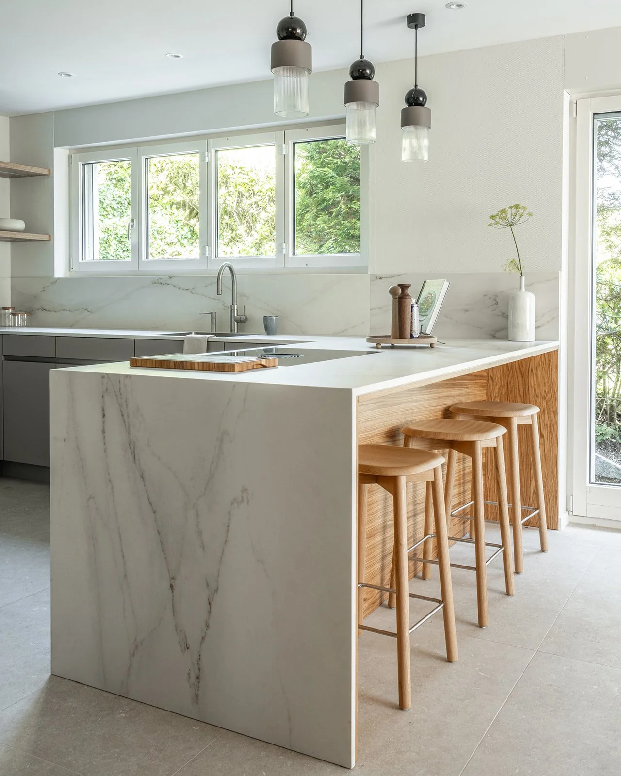 Kitchen island with waterfall stone worktop, warm oak cabinetry and wooden bar stools beside large garden windows.