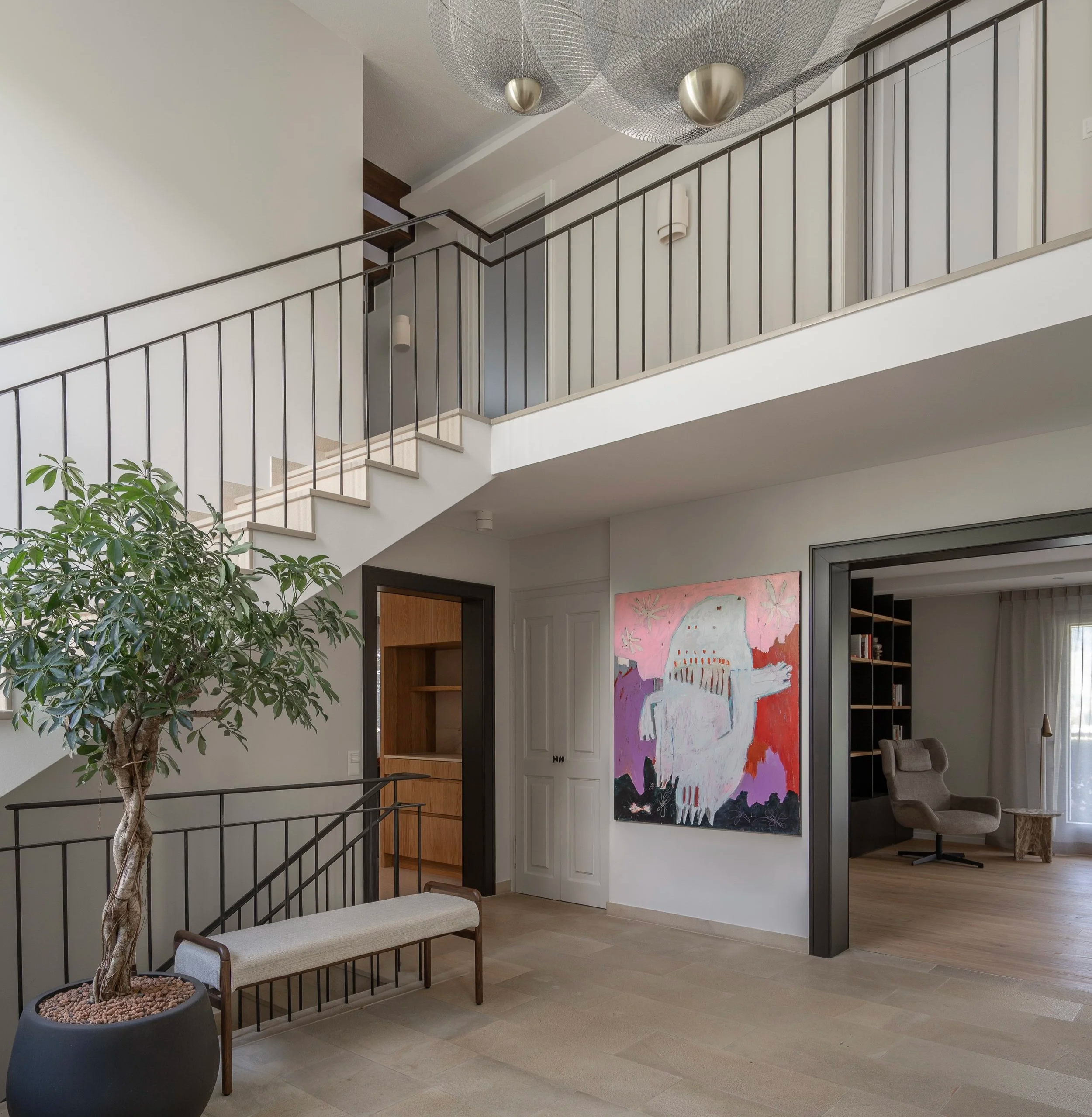 Double-height entrance hall with staircase, large artwork, indoor tree, bench and black metal railings.