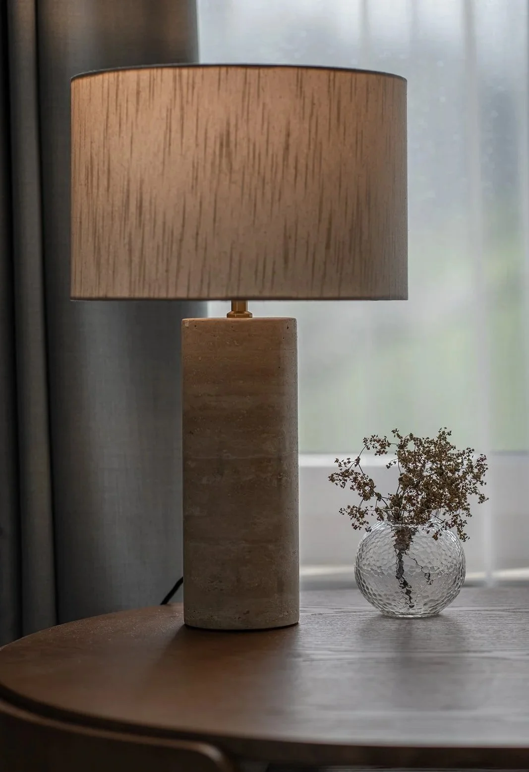 Detail of the desk with a stone table lamp, textured shade and small glass vase, combining warm natural materials.