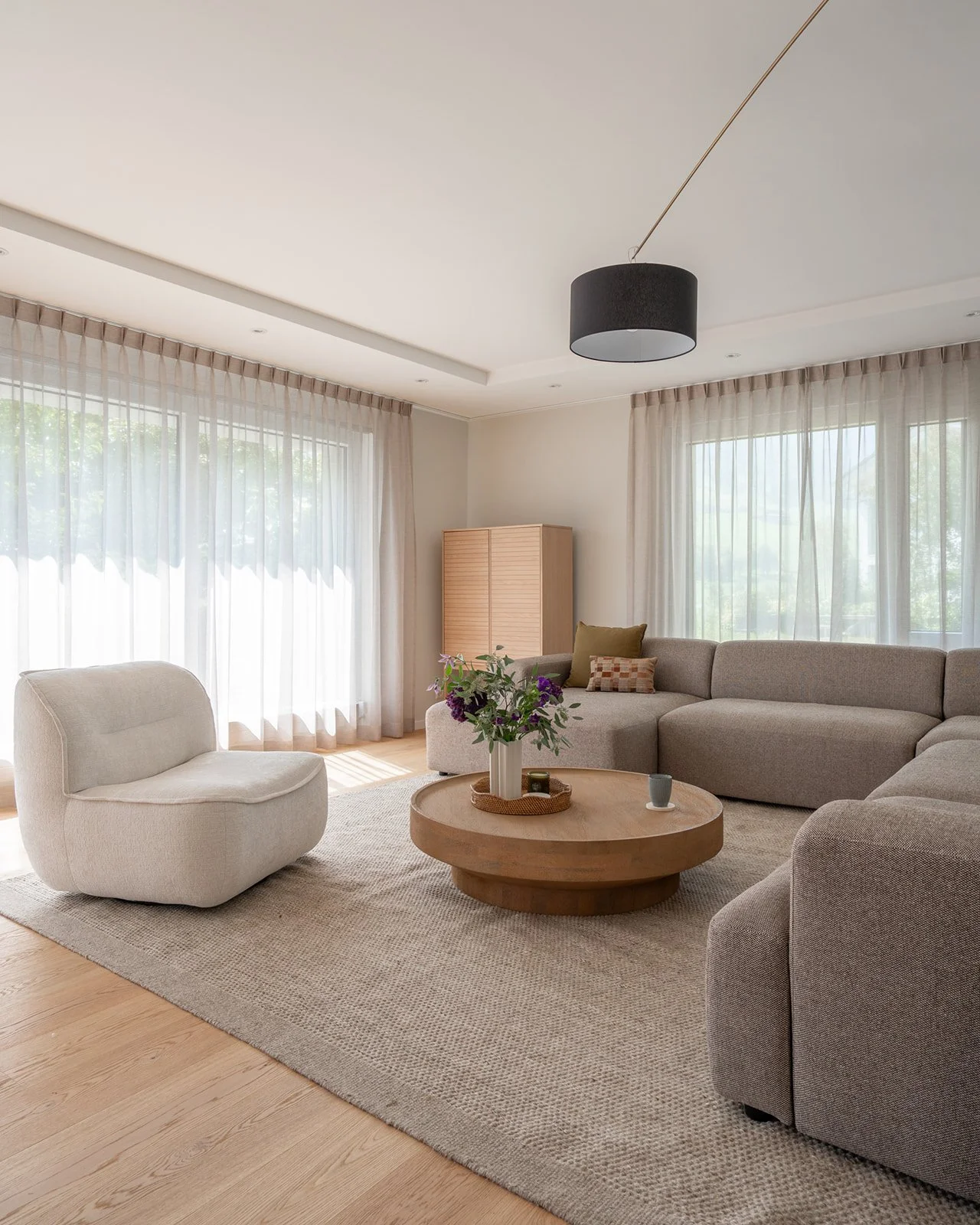 Bright living room with a soft-corner modular sofa, sculptural lounge chair, round wooden coffee table and architectural floor lamp at Landhaus Ennetmoos.