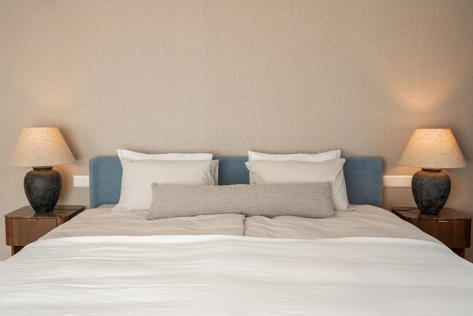 Front view of the master bed with linen-textured wallpaper, a soft blue upholstered headboard, layered white and sand-beige bedding, and linen bedside lamps on marble-topped tables.