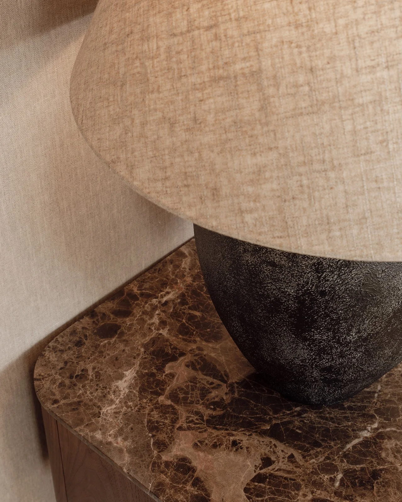 Detail of a bedside lamp with a textured dark base and linen shade on a brown marble bedside table.