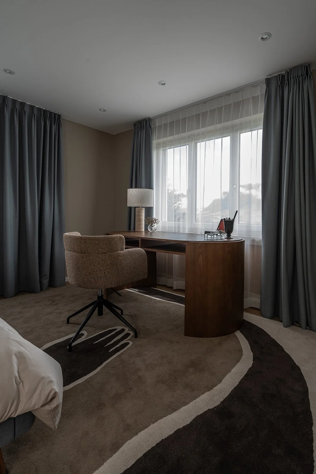 Guest room and home office with a sculptural wooden desk, upholstered swivel chair and graphical rug, framed by layered blue-grey curtains.
