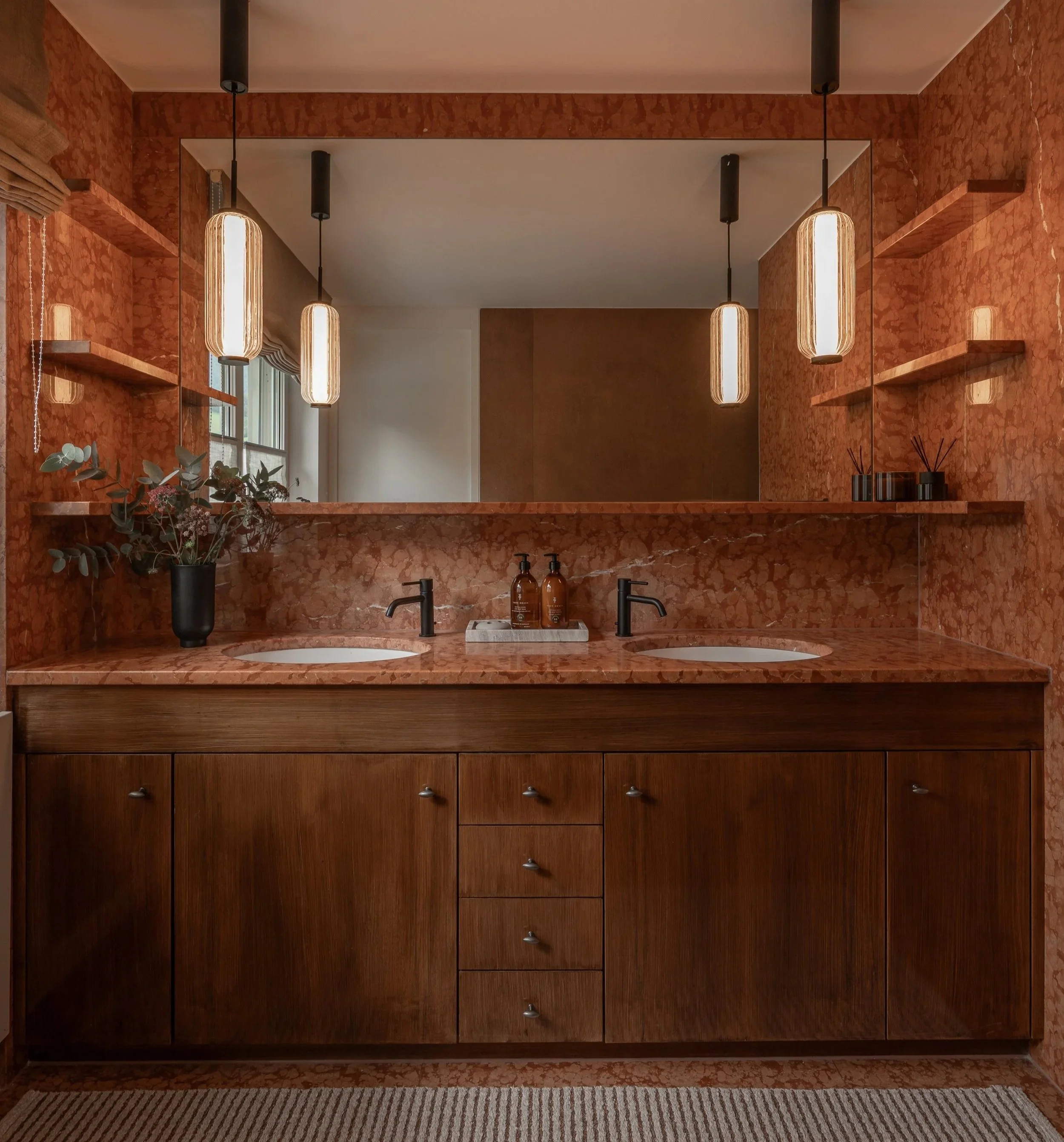 Master bathroom with a double vanity in warm wood, richly veined red marble surfaces, integrated shelving, a large wall mirror and ribbed glass pendant lights.