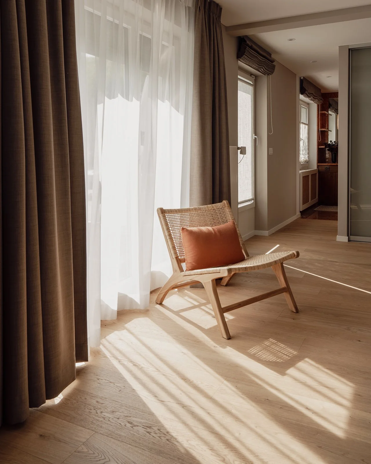 Sunlit corner of the master bedroom with a woven lounge chair positioned by the window for relaxing, reading and enjoying the view, framed by layered curtains and warm oak flooring.