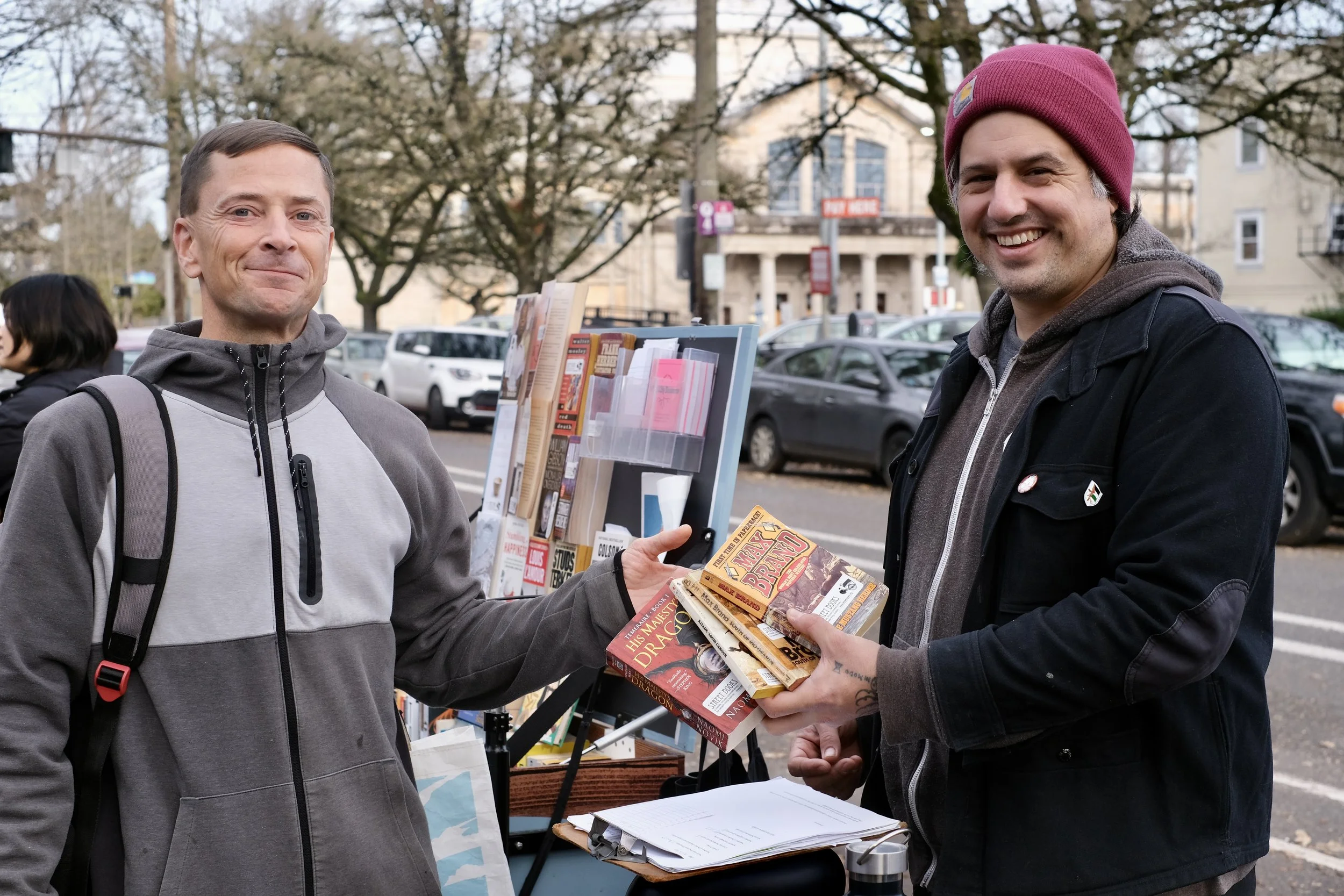 two smiling people dressed in winter clothes exchange a stack of paperback books in front of a mobile bicycle library on a city sidewalk