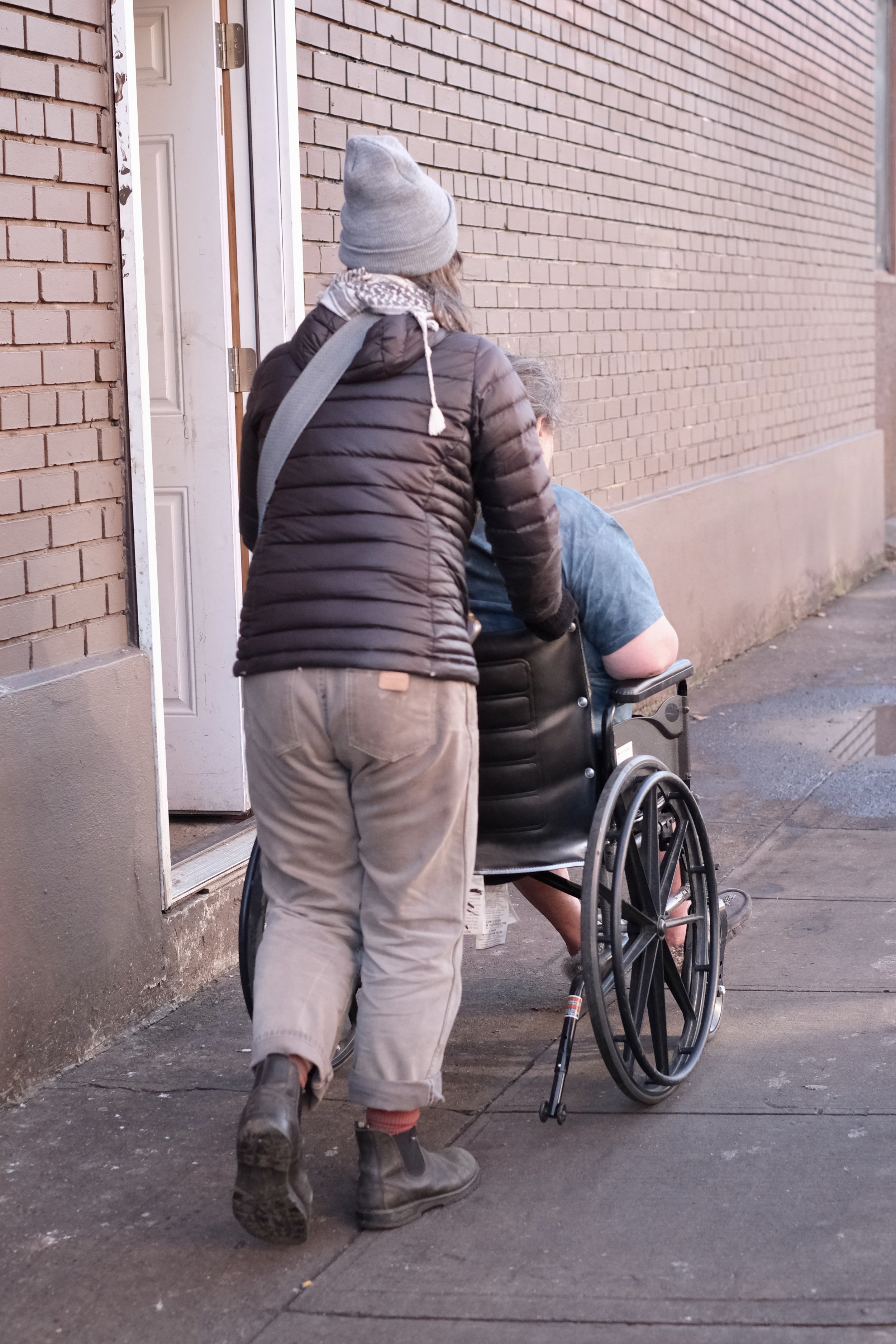 A person dressed in winter clothes pushes another person in a wheelchair down a city sidewalk in a photo taken from behind