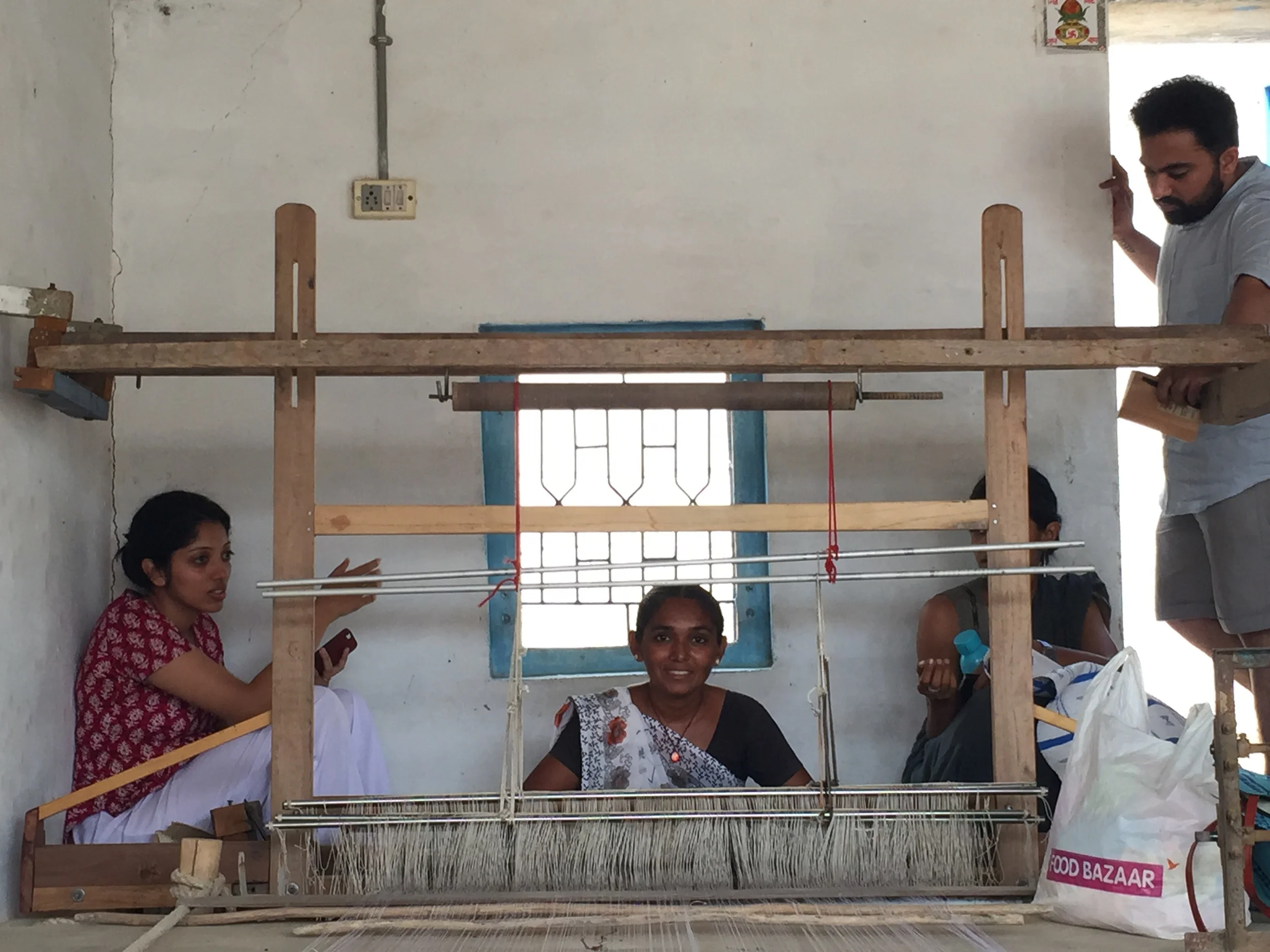 Arushi sits with Raji Ben and Shabri by the loom as Raji Ben creates a plastic weaving sample for The Initiative.
