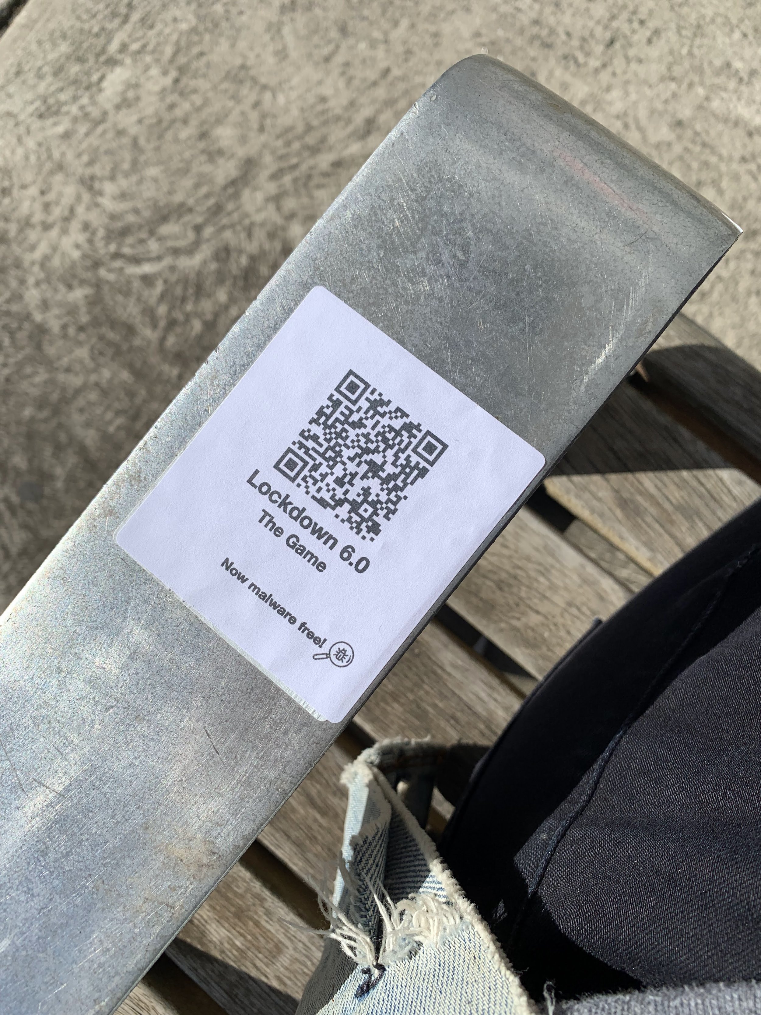  Alt text: A QR code on the arm of a park bench reads: Lockdown 6.0. The Game. Now malware free. 