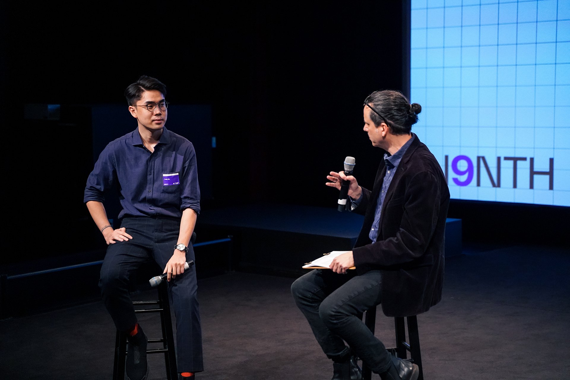  Student Felix Ho in conversation with Allan Chochinov. The two are sitting on stools and Allan is holding a microphone 