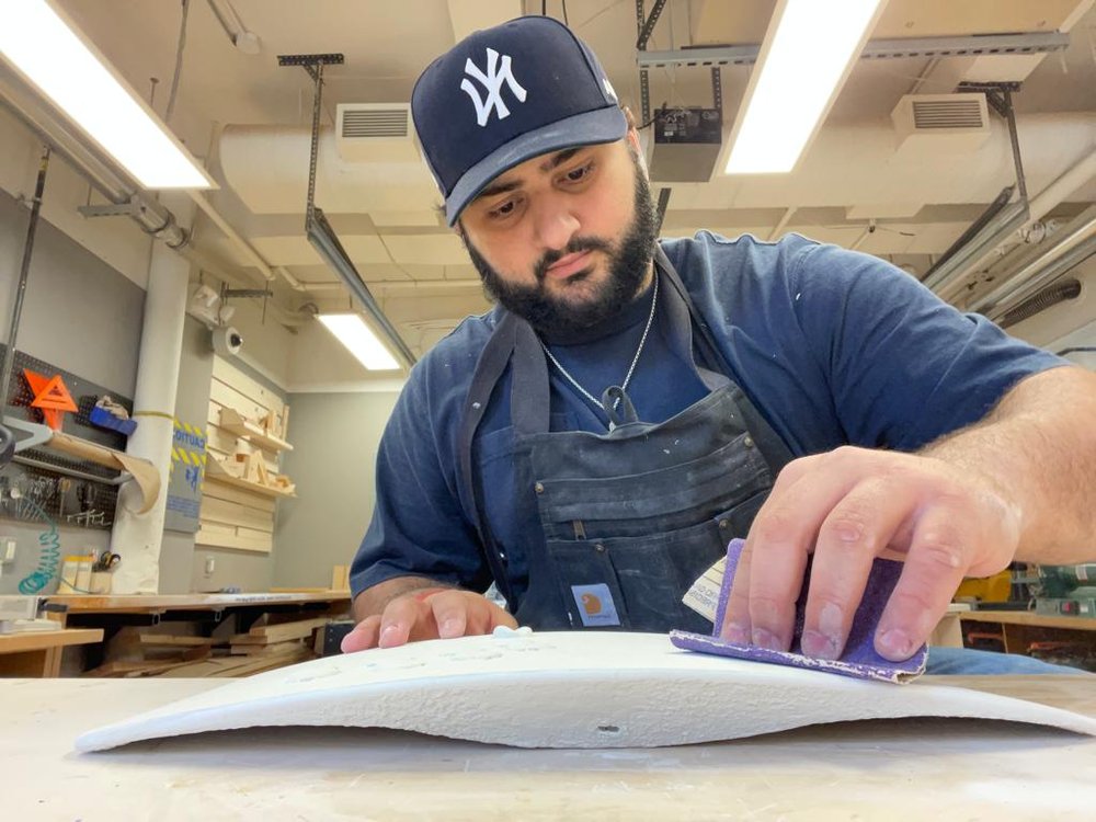  Image of student Jose Martin wearing a baseball cap and sanding a wooden prototype. 