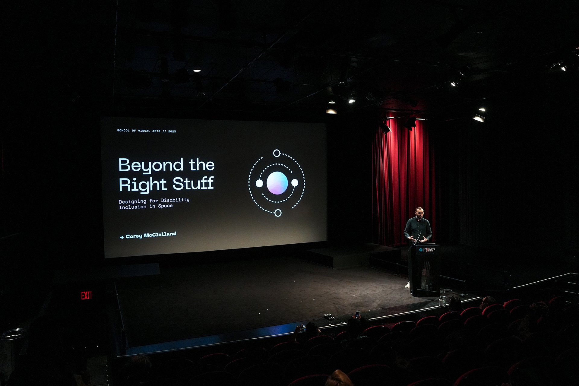  View of SVA Theatre stage at a distance with speaker at podium. The screen says “Beyond the Right Stuff” on a black graphic. 